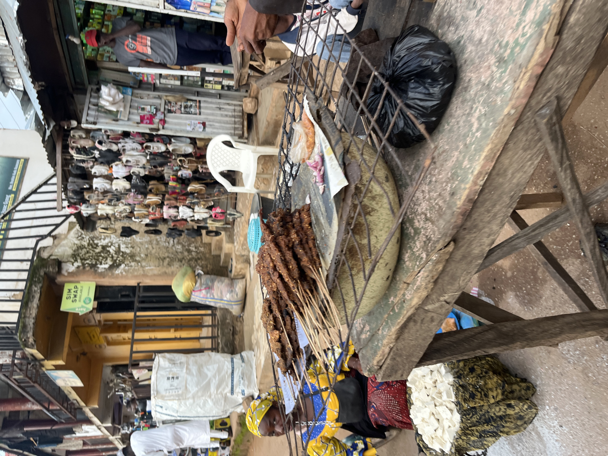 In Bukuru, Nigeria, a street scene unfolds with a food stall as the central focus. A woman, adorned in a yellow and blue patterned dress and headscarf, sits near a grill laden with skewers of grilled meat. In front of her, a mound of white, cubed food sits on a patterned cloth. The stall itself is rustic, constructed from weathered wood. To the right, a person's hands are visible, suggesting a transaction or interaction near another vendor. In the background, a variety of goods are displayed outside shops, including shoes and bags. A "SIM SWAP AVAILABLE" sign is prominently displayed on one of the buildings. The overall environment suggests a bustling market or street-side commercial area.