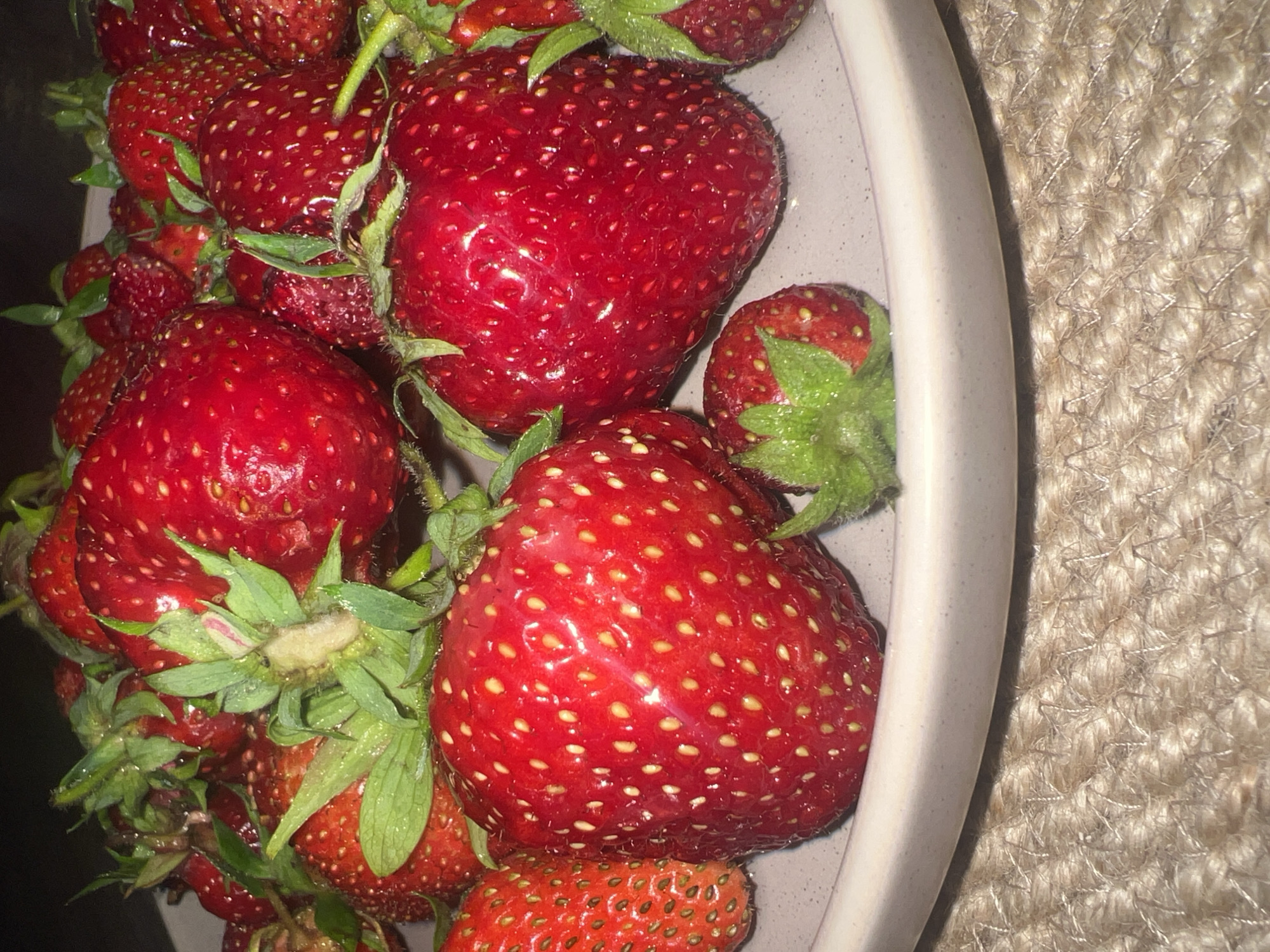 A close-up, high-angle shot captures a pile of ripe, red strawberries arranged on a light-colored plate. The strawberries vary in size, with some larger specimens dominating the foreground and midground. They are glistening and appear fresh, with visible seeds dotting their surfaces. Green stems and leaves are attached to most of the berries, adding a touch of natural color. The plate itself is positioned on a coarse, woven, light-brown surface, possibly a placemat or tabletop made of natural fibers. The background is dark and out of focus, drawing attention to the texture and vibrant color of the strawberries. There is no indication of human presence or activity in this image, nor any specific landmarks or environmental cues that would allow for pinpointing a location within Votkinsk, Russia. The image appears to be a still-life photograph of fruit.