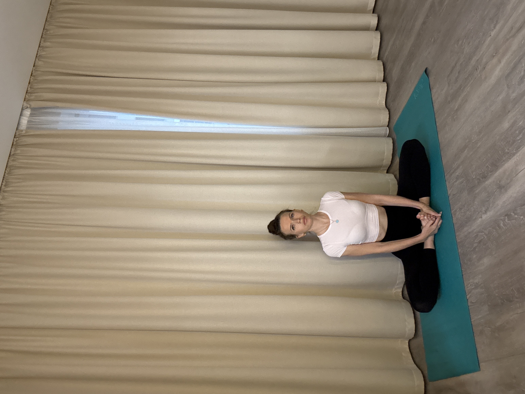 A woman is performing yoga on a turquoise mat in a room with light beige curtains. She is sitting in a cross-legged position with her feet pressed together, knees bent outwards, and hands clasped in front of her. She is wearing a white crop top and black leggings. Her hair is pulled back into a bun. The floor is made of grey wood-patterned laminate. There is a partially visible window with sheer white curtains behind the woman. The image appears to be taken indoors. There is no information in the image to suggest the location is in Dolgoprudny, Russia.