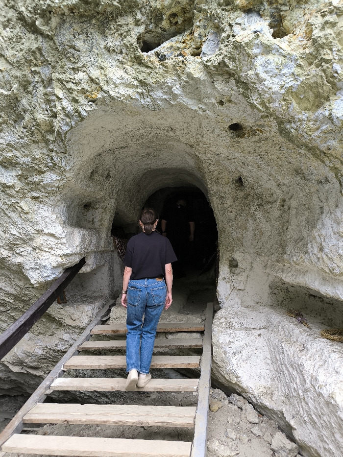 The image depicts the entrance to a man-made or natural cave system in Horaivka, Ukraine, carved into a light-colored rock formation. A single individual, viewed from behind, is ascending a set of wooden stairs leading into a dark, arched tunnel entrance. This person is wearing a black short-sleeved t-shirt, blue denim jeans, and light-colored closed-toe shoes. Their hair is dark and pulled back into a ponytail.
The stairs consist of rough-hewn wooden planks supported by a metal frame, with a dark metal handrail extending along the left side. The rock face surrounding the entrance is a textured, light grey to off-white material, possibly limestone or chalk, exhibiting irregular patterns and small, dark apertures in its upper sections. The interior of the tunnel beyond the immediate entrance is dark, with the faint outline of at least one additional figure discernible deeper within the gloom. The ground immediately around the base of the stairs consists of loose soil and rock debris. No direct interactions between individuals are visible. The environment appears to be a natural rock formation modified for access, indicative of a historical or natural site.