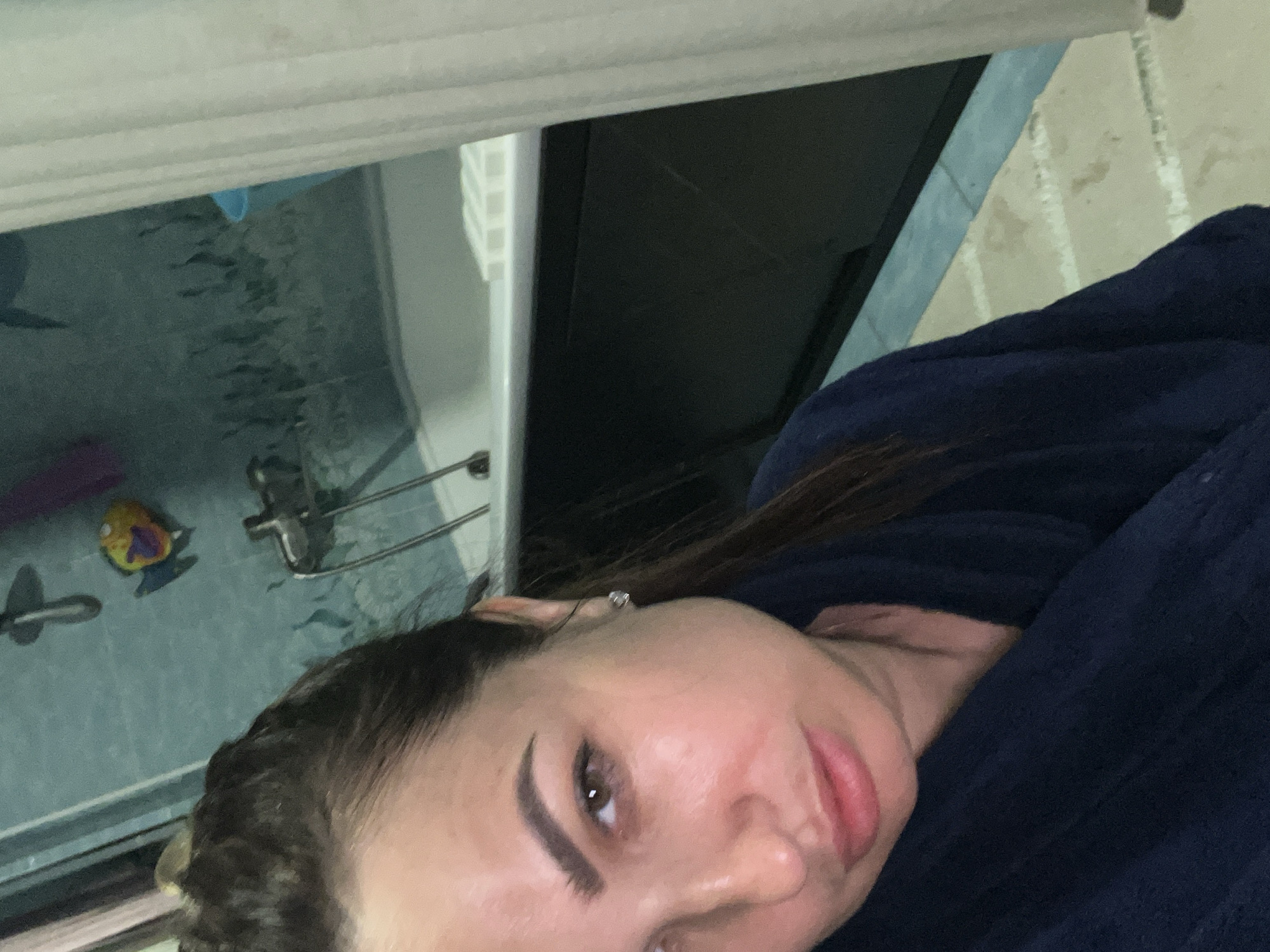 A woman, with brown hair pulled back and wearing a dark blue robe, is pictured in what appears to be a bathroom. She is looking at the camera with a neutral expression. In the background, a bathtub is visible with a shower head and a colorful bath toy. The walls are tiled in a light blue color, and a white curtain is present to the right of the frame. The floor appears to be tiled in a beige and white pattern.