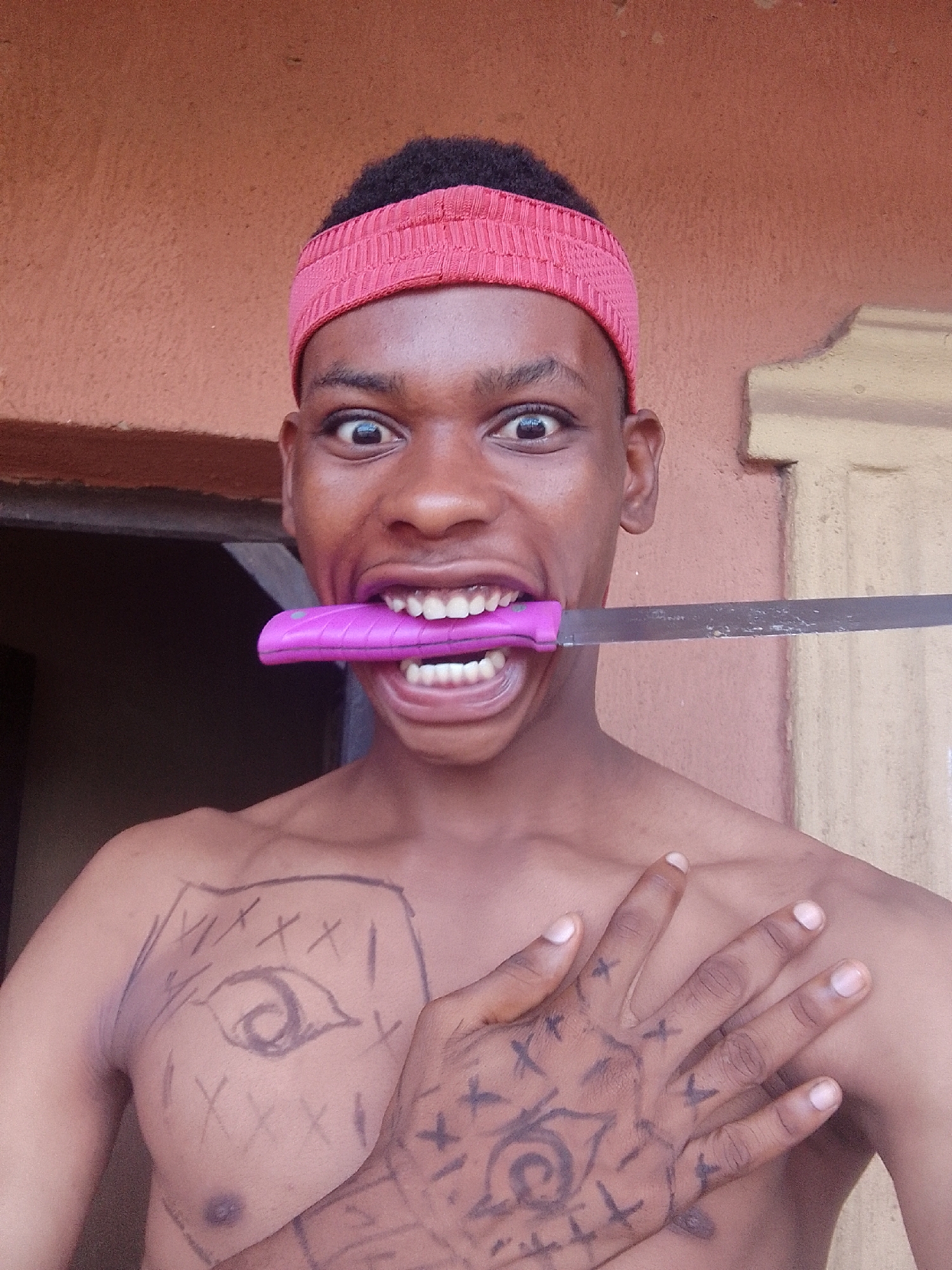 The image captures a young person, likely male, with dark skin and short, dark, curly hair, wearing a red, textured fabric headband. He is shirtless, presenting an intense expression with wide-open eyes and an agape mouth, gripping a knife horizontally between his teeth. The knife has a bright pink handle and a silver, somewhat dull and scratched blade.
On the subject's chest, dark marker drawings are visible. A prominent eye symbol, enclosed within a bordered shape and surrounded by several "X" marks, is drawn on the left side of his chest (viewer's right). His right hand (viewer's left) rests over his chest, partly covering this drawing. This hand also features numerous dark marker drawings, including a circular symbol resembling a spiral or eye on the palm, and various "X" marks and other small symbols scattered across his fingers and the back of his hand.
The background behind the person consists of a textured, reddish-brown wall, possibly stucco or plaster. To the right, a darker opening, perhaps a doorway or corridor, is visible. The lighting suggests daytime conditions, casting a relatively even light on the subject. The overall mood conveyed by the subject's expression and the knife is striking and dramatic. There is no visible legible text in the image.