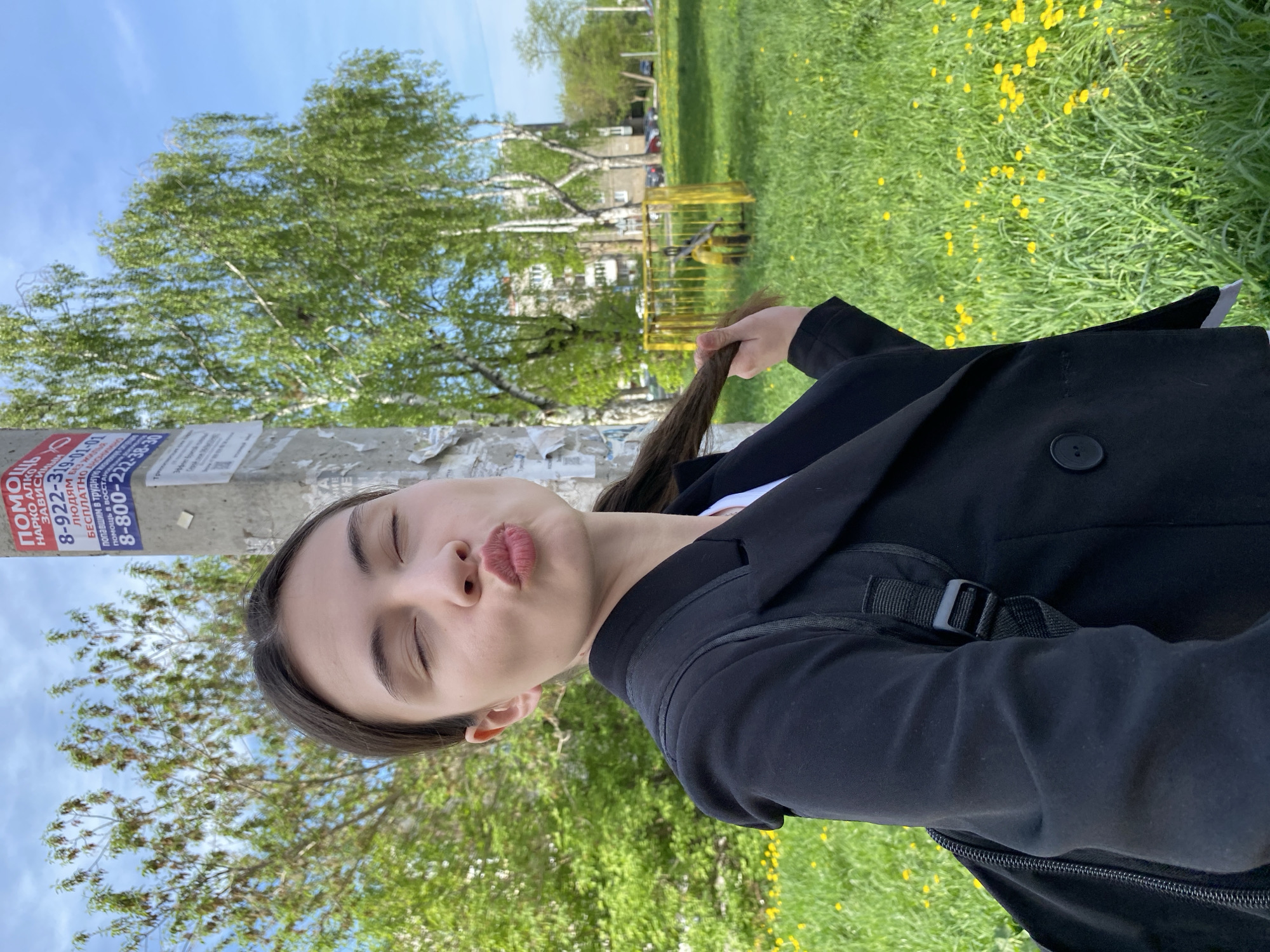 The image shows a woman outdoors, standing next to a utility pole plastered with advertisements, one displaying Russian text and phone numbers that suggest help with addiction. She is wearing a black blazer over a white shirt, and a black backpack. She is puckering her lips into a kissing face with her eyes closed and holding a section of her dark hair in her hand. The background features a green grassy area with yellow flowers, likely dandelions, with trees, a yellow playground structure, and buildings visible in the distance. Given the text on the advertisements, this image is likely in Votkinsk, Russia.