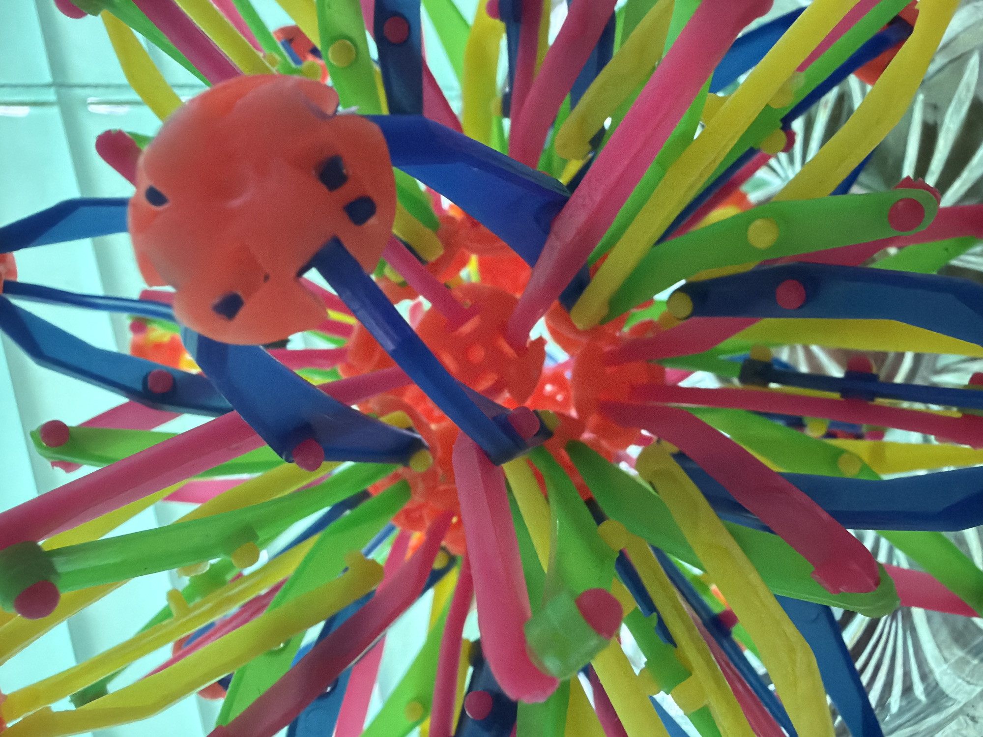The image shows a close-up view of a colorful Koosh ball toy. The toy is composed of numerous flexible, narrow strands in a variety of colors including pink, yellow, green, and blue, all radiating outward from a central, orange, perforated rubber core. Small cylindrical caps, with colored tops matching the strands' colors, are fixed to the ends of many strands. The background appears to be a blurry, light-colored surface, possibly a wall. The photo appears to have been taken looking upwards towards the toy. The image does not contain any information to determine if it was taken in Bugulma, Russia.