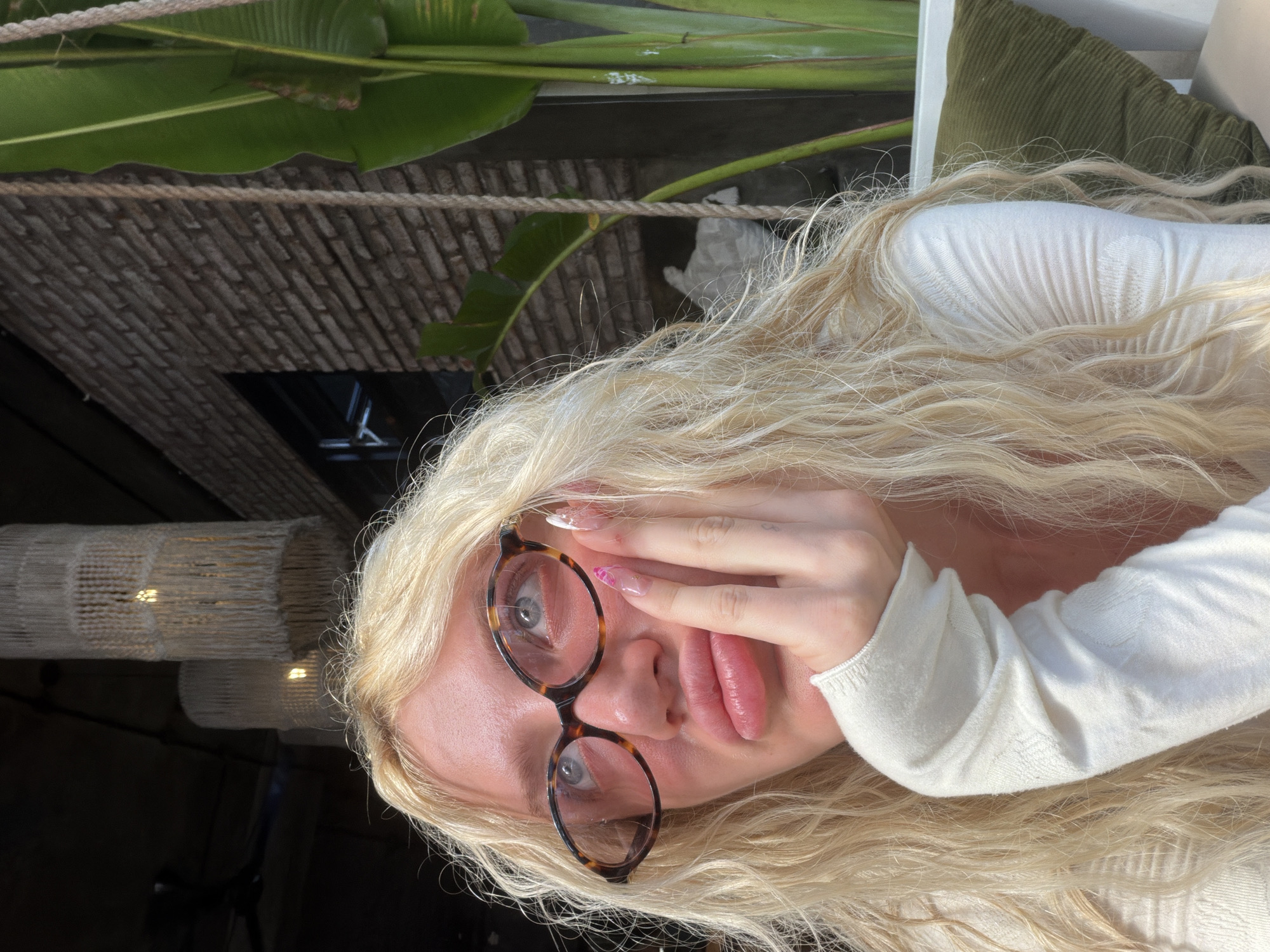 A woman with long, blonde, wavy hair and light-colored eyes is captured from a medium close-up angle. She is wearing large, tortoise-shell glasses and a cream-colored long-sleeved top. Her left hand is raised, with fingers resting on her chin and cheek, while her lips are pursed. Her nails are long and painted with a light pink and white design. She is looking upwards and to the left, with her head tilted back slightly.
The background suggests an outdoor or semi-outdoor setting, possibly a restaurant or cafe in North Kuta, Indonesia. Behind her, a brick wall is visible, along with large, green leaves of a tropical plant. Hanging from the ceiling are decorative lamps with a woven, fringe-like appearance. The lighting indicates it is daytime, with sunlight casting shadows. A green, corduroy pillow is visible in the lower right corner, next to a white chair.