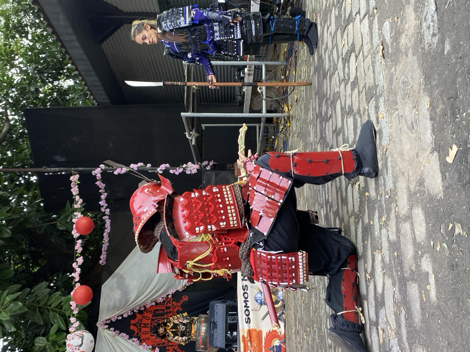 An outdoor cultural event is depicted in a public space in Buenos Aires, Argentina, identifiable by its cobblestone paving and mature trees. Two individuals in elaborate samurai armor are positioned on the ground.
On the left, a person kneels in a full suit of red samurai armor, including a helmet, chest plate, shoulder guards, thigh guards, and shin guards, all adorned with intricate white and gold detailing. Black undergarments and tabi-style footwear are visible. A katana in a red scabbard is sheathed at their left hip. The individual faces generally right, with their back partially towards the viewer, in a low, crouched posture.
On the right, a female individual stands upright, facing the kneeling figure. She wears dark blue and black samurai-style armor, also featuring chest, shoulder, arm, and leg protection, with light accents. Her hair is pulled back, and her facial features are visible. She holds a naginata, a polearm with a curved blade, vertically in her right hand, with the butt end resting on the ground.
In the background, a temporary stage setup features a black backdrop, white fabric drapes, and festive decorations including red paper lanterns and strings of pink artificial cherry blossoms. A banner partially visible on the stage reads "SOMOS N..." with a smaller banner below displaying a Viking helmet and Japanese-style imagery of a dragon and waves. Speakers and other stage equipment are discernible. A building with a corrugated metal roof is partially visible to the far right. The scene suggests a performance or ceremonial demonstration.