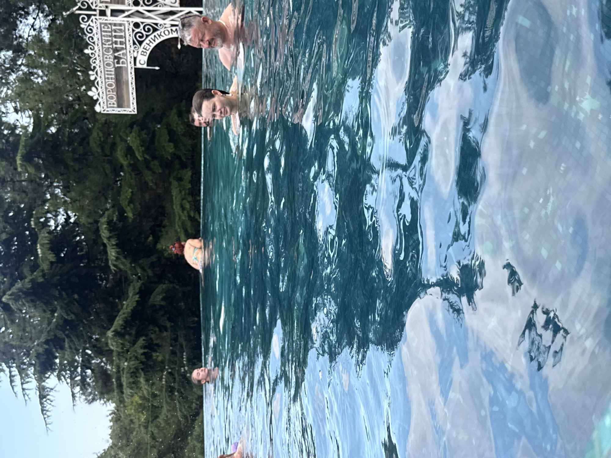 In the city of Yalta, Ukraine, an outdoor pool with clear, deep blue water is filled with people enjoying a swim. A sign with Cyrillic lettering, "ВОРОНЦОВСКИЕ БАНИ" (Vorontsovskie Baths), is visible in the background, attached to an ornate white structure. Tall, dark green evergreen trees surround the pool, creating a natural and lush setting. The water's surface reflects the sky and trees, creating a dynamic visual effect. Several individuals are visible in the water, with their upper bodies submerged, suggesting a relaxed and recreational atmosphere. A man with a grey beard and sunglasses on his head is in the foreground, facing right, while a woman with dark hair is to his left. Another woman with reddish hair is further back on the left side of the frame, wearing a light-colored bikini. The overall scene captures a moment of leisure and enjoyment in an outdoor pool setting within a scenic environment.
