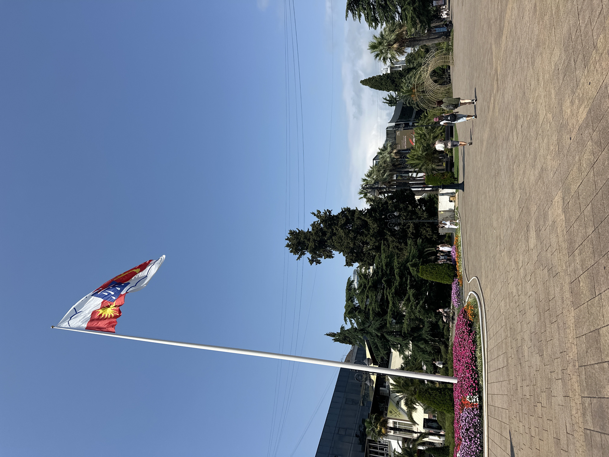 In the city of Sochi, Russia, a large flag with red, white, and blue elements, including a sun emblem, flies atop a tall white pole in a paved public square. The square features a circular flower bed filled with pink and purple flowers, accented with orange and red blooms. Several people are visible in the distance, walking across the square. Behind the flower bed, various trees, including palm trees and tall evergreen trees, are present, along with buildings that appear to be shops or restaurants. The sky above is clear and blue.