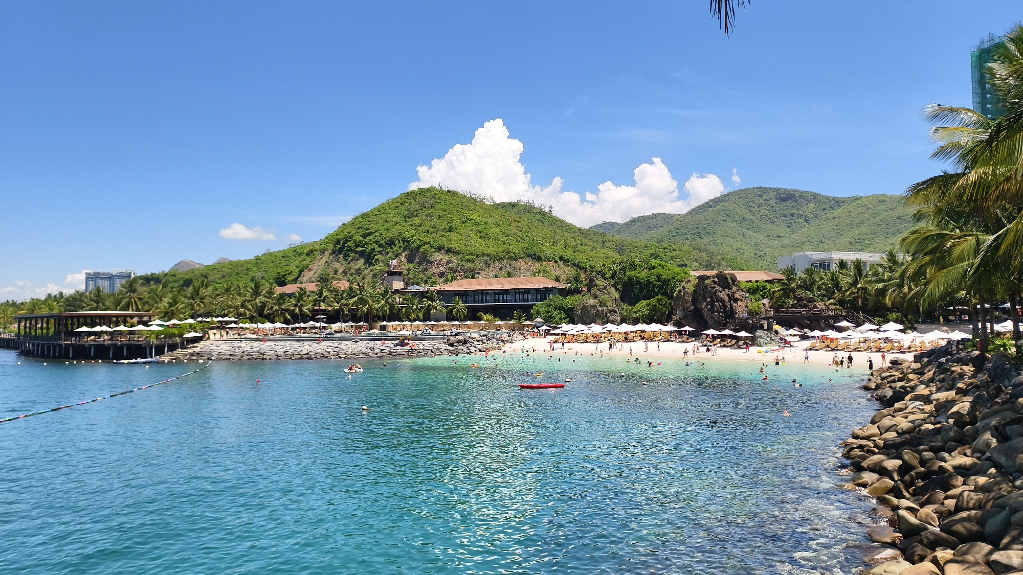 This is a panoramic, eye-level, medium shot of a tropical beach in Nha Trang Ward, Vietnam. The image features a clear, turquoise sea in the foreground, with a sandy beach lined with white umbrellas and lounge chairs in the middle ground. A lush, green hill dominates the background, with a large, multi-story building nestled into its side. Several people are visible on the beach and in the water, swimming and sunbathing. A small, red kayak is floating in the water. The sky is a vibrant blue, with scattered white clouds. The scene is bathed in bright sunlight, suggesting a warm, sunny day. The overall impression is one of a peaceful and idyllic vacation destination.