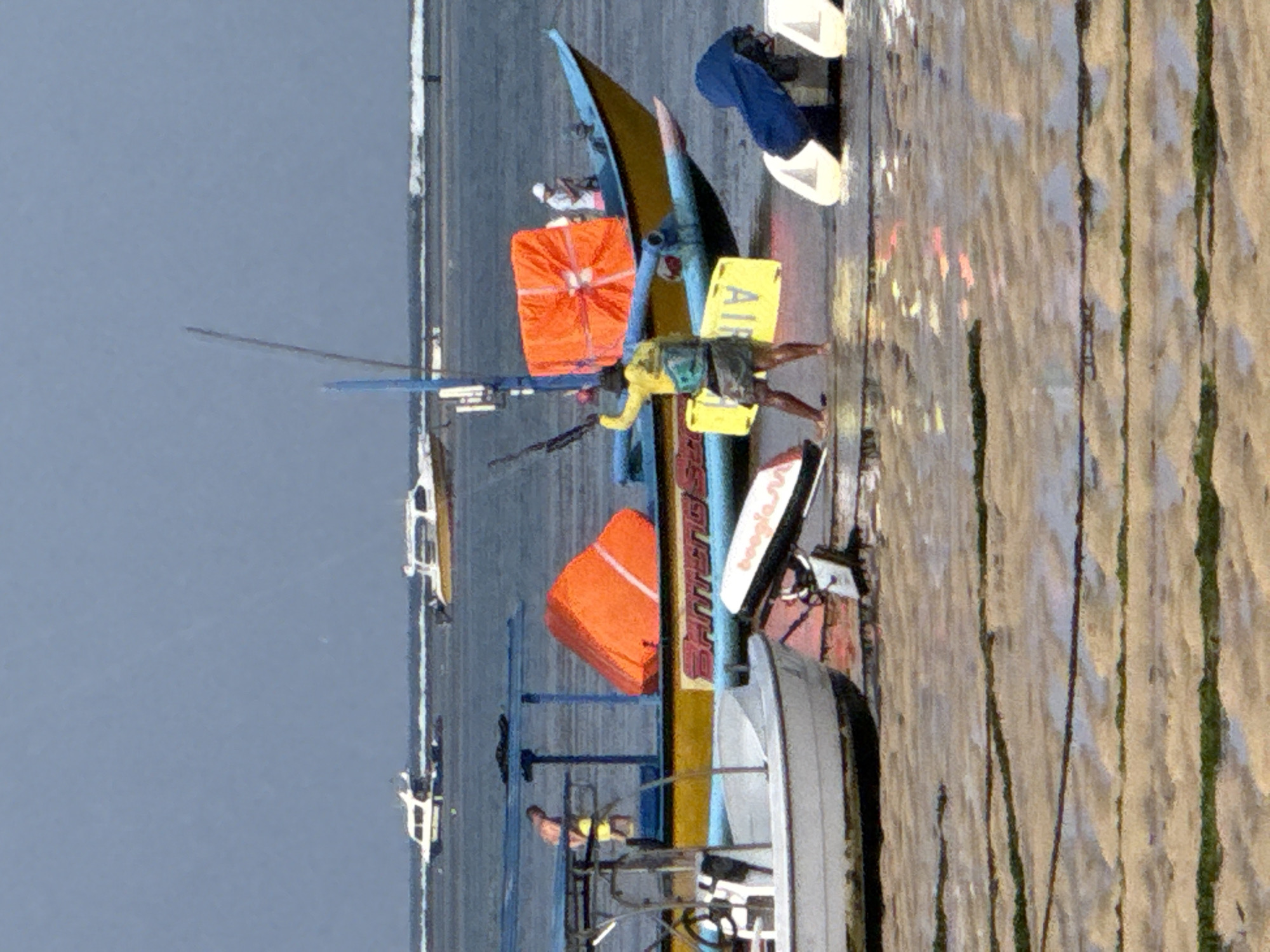 The image depicts a waterside scene in Denpasar, Indonesia, likely at a beach or harbor. In the foreground, the wet sand is partially covered with strings of seaweed. Several boats are visible. The most prominent boat is a yellow and blue vessel with an orange flotation device on its deck. A person in a yellow shirt and shorts is standing on the sand near this boat, holding what appears to be a rifle or long pole. Another person is visible on the deck of the yellow and blue boat, wearing a white hat. To the left of this boat, a white boat is partially visible. In the background, there are other boats, including white speedboats, on the water under a hazy sky. The water appears calm, with gentle waves.