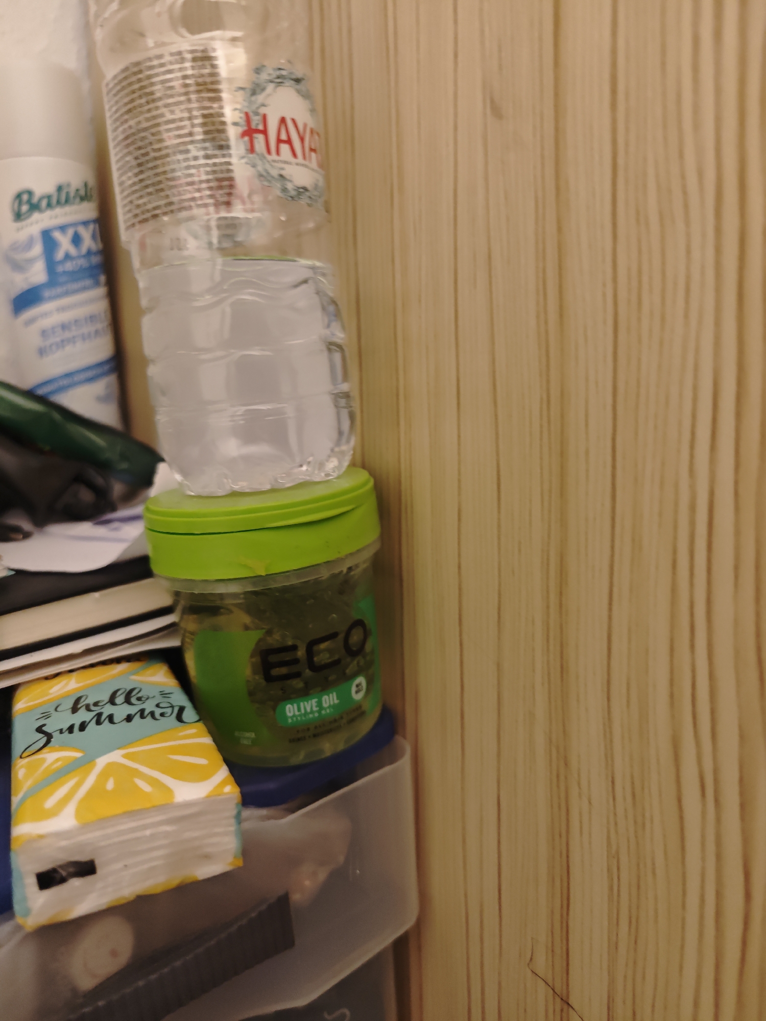 In a dimly lit indoor setting, possibly a bathroom or bedroom, several items are stacked on a shelf against a wood-paneled wall. The objects include a partially filled plastic water bottle, a green jar of hair gel labeled "ECO OLIVE OIL," and a white bottle with blue lettering that reads "Batiste XXL." Below these items, a stack of books or notebooks is visible, with a decorative item featuring lemons and the words "hello Summer" on top. To the right, a transparent plastic container holds more items, partially obscured by the other objects. The scene appears to be a personal collection of toiletries and accessories. The location is not specified as Zweibrücken, Germany.