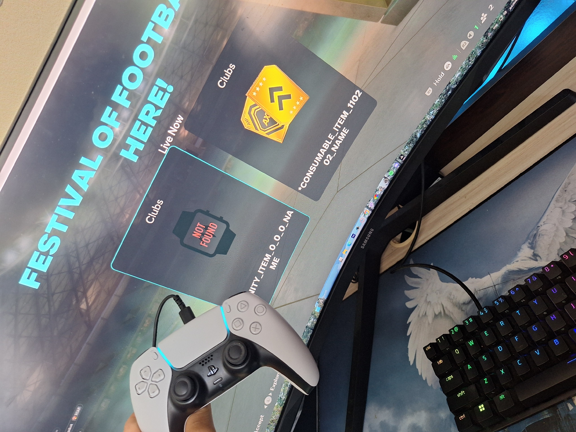 The image displays a close-up, high-angle shot of a gaming setup. In the foreground, a white PlayStation 5 DualSense controller is held by an unseen hand, with its characteristic illuminated accents visible. To the right, a black mechanical keyboard with RGB backlighting is positioned. The primary focus is on a large computer monitor displaying a game interface, specifically from what appears to be a sports video game given the "FESTIVAL OF FOOTBALL HERE!" text and the "Clubs" section visible on screen. One card on the screen shows a "NOT FOUND" message overlaid on a watch icon, while another displays two golden ticket-like items with symbols and star ratings, labeled as "*CONSUMABLE_ITEM_1102_02_NAME". The background of the monitor shows a blurred stadium or arena. The overall environment suggests a modern indoor space dedicated to gaming. There is no visible indication of the city of Varna, Bulgaria, nor any details that could place the scene in a specific geographical location.
