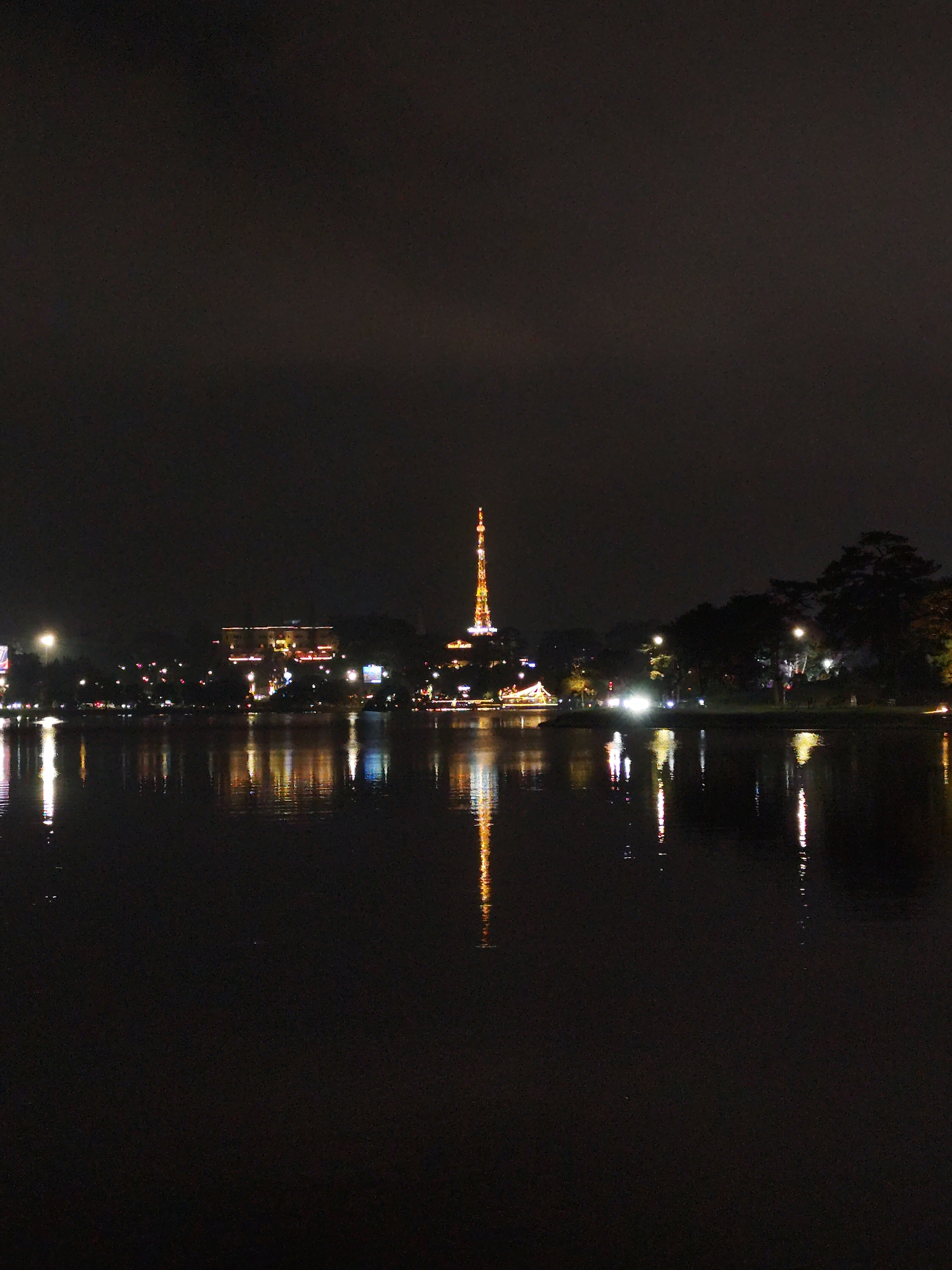 A nighttime cityscape reflects off a calm body of water. Dominating the skyline is a tall, illuminated tower, likely a communications or observation structure. The water's surface mirrors the lights from the tower and other buildings along the shore. The scene appears to be in Da Lat, Vietnam, identifiable by the tower's distinctive architecture. The general environment suggests a serene and possibly tourist-oriented location. The image does not show any individuals.