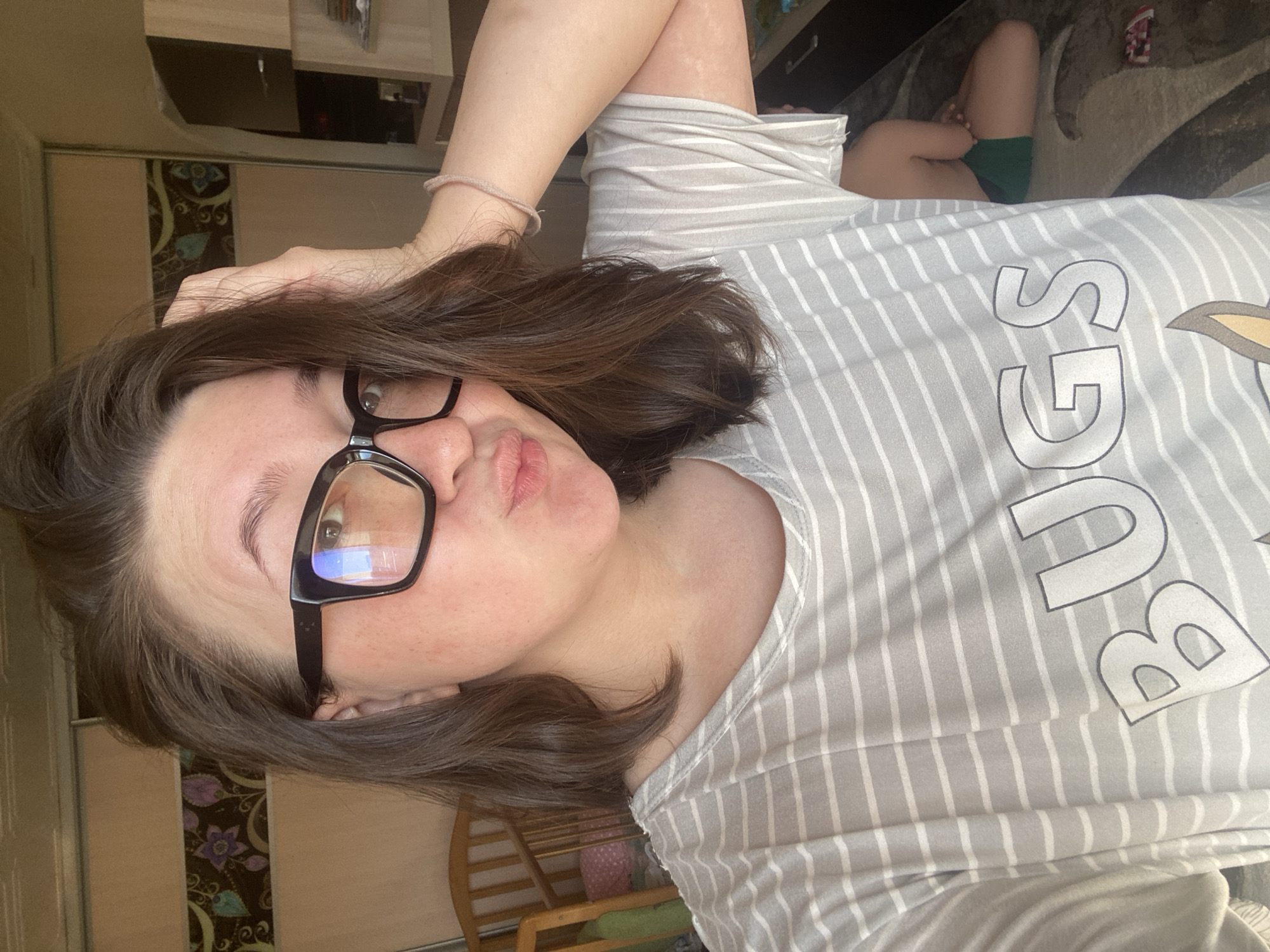 A woman with short brown hair and glasses is posing for a selfie. She is wearing a light gray striped t-shirt with the word "BUGS" printed on it. The location appears to be indoors, with a closet and wooden furniture visible in the background. The exact city of Chaikovsky, Russia, cannot be confirmed from the image alone.