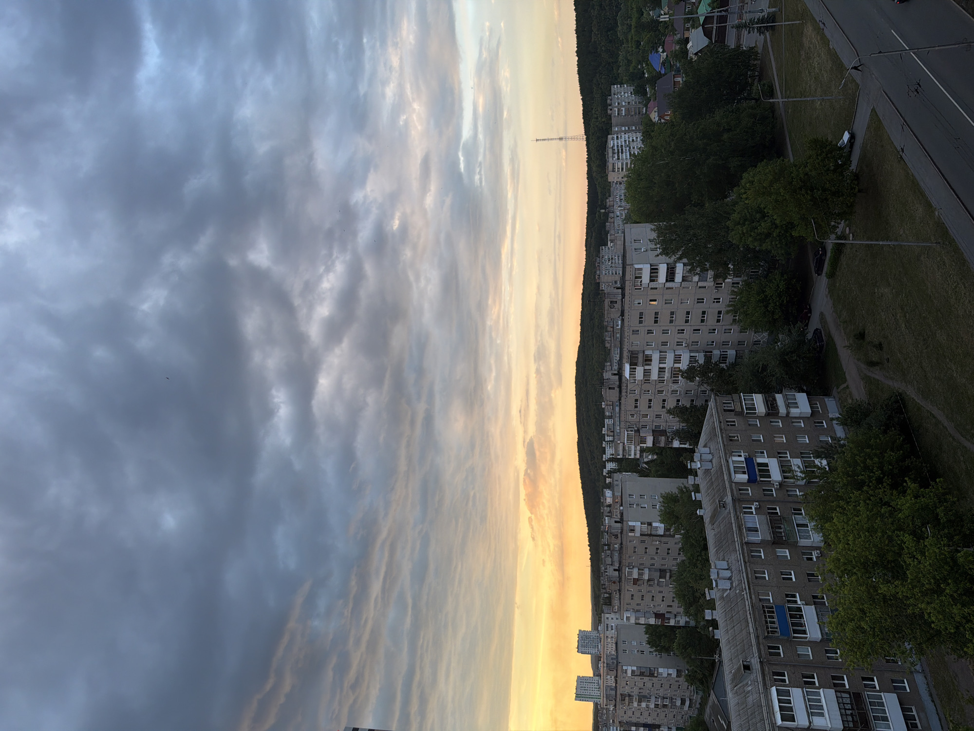 This media file captures an urban landscape at either sunrise or sunset, with the sky transitioning dramatically from dark, overcast clouds on the left to warm, orange and yellow hues on the right, where the sun's light is breaking through. The image is oriented vertically, but depicts a horizontal panorama when viewed correctly.
The scene is dominated by numerous multi-story residential buildings, likely apartment blocks, characterized by their uniform, often grey concrete facades, multiple windows, and balconies. These buildings appear typical of Eastern European or post-Soviet architecture. Many trees and patches of green space are interspersed among the buildings, and a dense line of forest or hills runs along the horizon, separating the urban area from the distant sky.
A prominent, slender antenna or transmission tower stands on the horizon, silhouetted against the bright part of the sky. To the far right, a road or street with a white dividing line is visible, suggesting traffic infrastructure. A few streetlights or utility poles are also scattered throughout the scene. Although no explicit human activity is depicted, the urban environment suggests daily life within the buildings. Small specks, possibly birds, can be seen against the cloudy sky.
The notable details include the striking contrast in the sky's colors, indicating the specific time of day. The cloudy weather adds texture and depth to the sky. No visible text is present in the image.