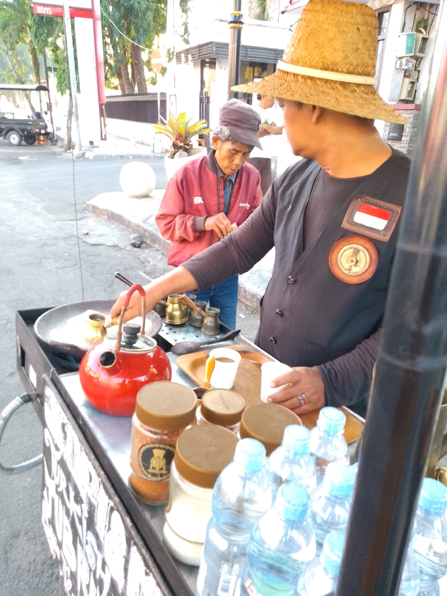 The image captures an outdoor street scene in Jember, Indonesia, focusing on a coffee cart. The cart is laden with various items, including a bright red kettle, brass coffee pots, jars of what appears to be coffee and sugar, and several plastic bottles of water. In the foreground, a person wearing a straw hat and a dark vest is preparing a beverage, with their hand reaching towards a brass coffee pot placed over a heat source. Another individual in a red jacket and a gray cap stands behind the cart, observing the process. The background reveals a street with trees, a building with an ATM sign, and a parked vehicle. The overall atmosphere suggests a busy street corner in a local Indonesian city.