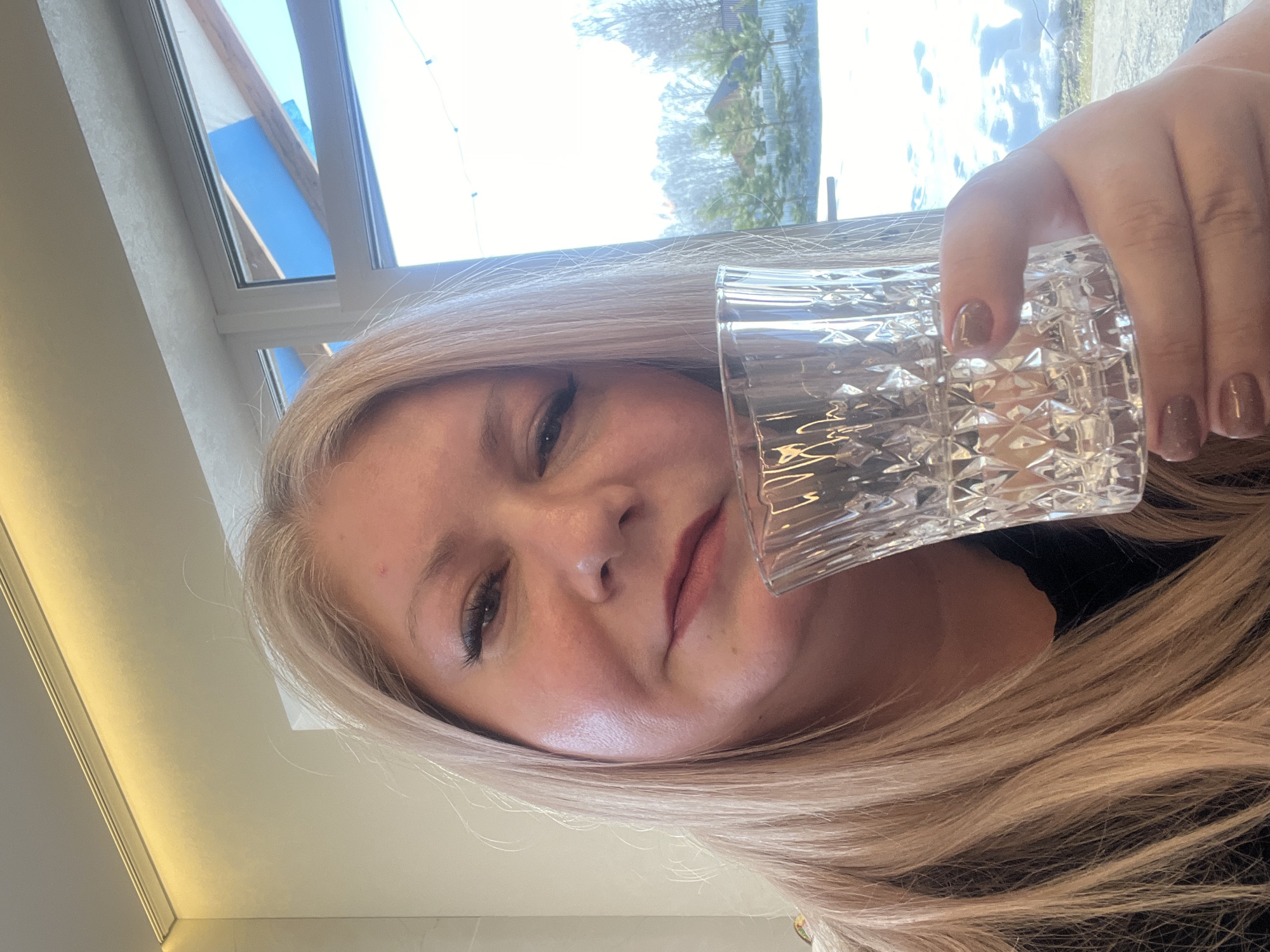 A middle-aged Caucasian woman with long, straight, blonde hair and dark eye makeup looks directly at the camera with a slight smile. She holds a clear, faceted glass containing a small amount of liquid in her left hand, which has light brown manicured nails. She is positioned indoors against a light-colored wall with a prominent white window frame to her right. Through the window, an outdoor scene is visible, characterized by snow-covered ground, several small coniferous trees, and bare deciduous trees in the mid-distance. A building with a dark roof and a light-colored fence are also present. The sky is partially visible and blue, with an architectural element displaying blue and wood tones overhead. This scene is situated within the city of Новая Чернушка, Russia.