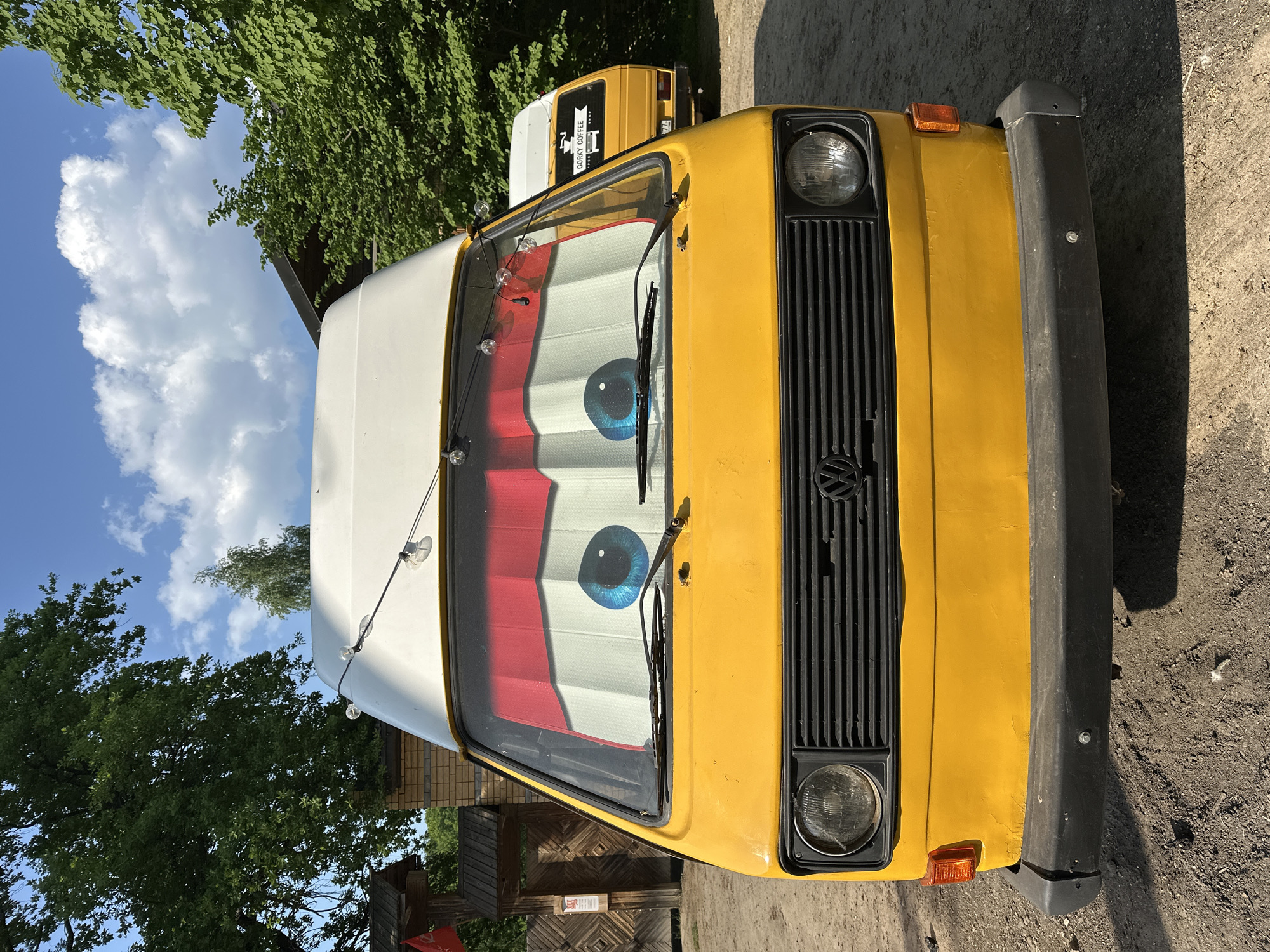 The image shows the front of a yellow and white Volkswagen van, with a pair of cartoonish blue eyes displayed inside the windshield. The van is parked on a dirt or gravel surface. Behind it, another similar van can be partially seen. Trees and a blue sky with clouds are visible in the background. The scene appears to be outdoors, likely in a parking area or a street setting. The presence of the Cyrillic-scripted 'GORKY COFFEE' sign on the van implies the location is Nizhny Novgorod, Russia.