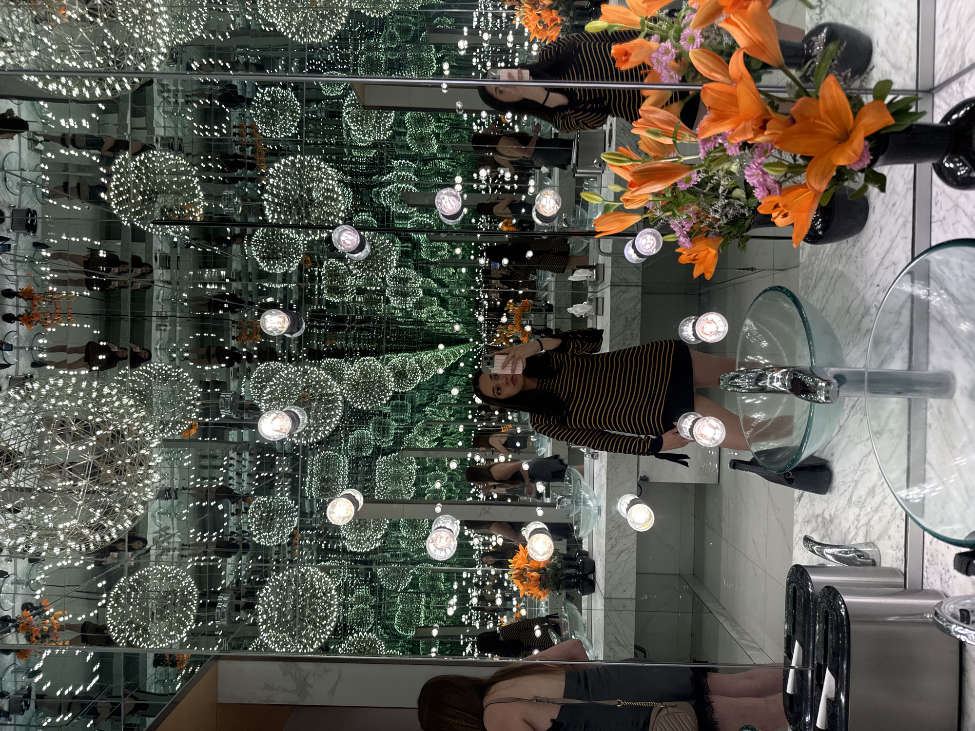 A person is taking a selfie in an infinity mirror room. The room contains hanging spherical lights and vertical mirrors, creating the illusion of a never-ending space filled with lights and reflections. A marble sink with a clear glass bowl and chrome faucet is visible. Orange and purple flowers are placed in a black vase. The location appears to be a restroom or vanity area due to the presence of the sink and grooming items. A second person is partially seen in one of the mirrors in the lower section of the image. The scene is set in a public or semi-public location, but the specific location within Puebla, Mexico, cannot be determined from the visual information provided.