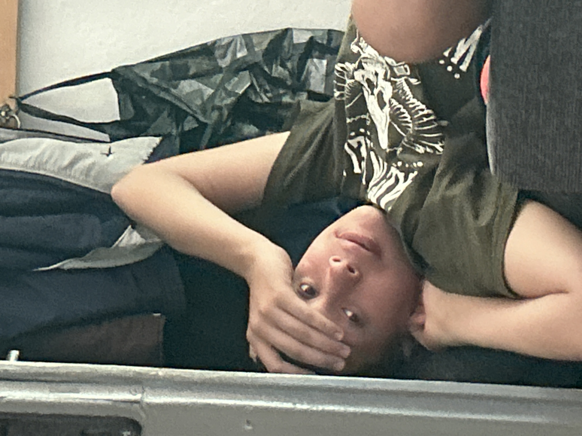 The media file shows a person lying on their back, with their head resting on a surface. Their body is oriented upside down relative to the viewer's perspective. The person is wearing a dark green t-shirt with a white graphic print on it. Their right hand is covering their mouth and nose, and their left arm is bent at the elbow, resting near their head. The person's eyes are open and looking towards the camera. The background consists of stacked fabric items, including what appears to be a dark grey blanket or sleeping bag and a lighter, crinkled material that could be part of a backpack or tarp. A metal rail is visible in the foreground, suggesting the person is on some form of elevated platform or bunk bed, possibly in a dormitory or camping setting. The lighting appears to be indoor, with no clear indication of time of day or weather conditions. The overall mood conveyed by the image is one of casual repose or perhaps slight discomfort due to the hand placement. No text is visible in the image. The scene is set in Govedare, Bulgaria.