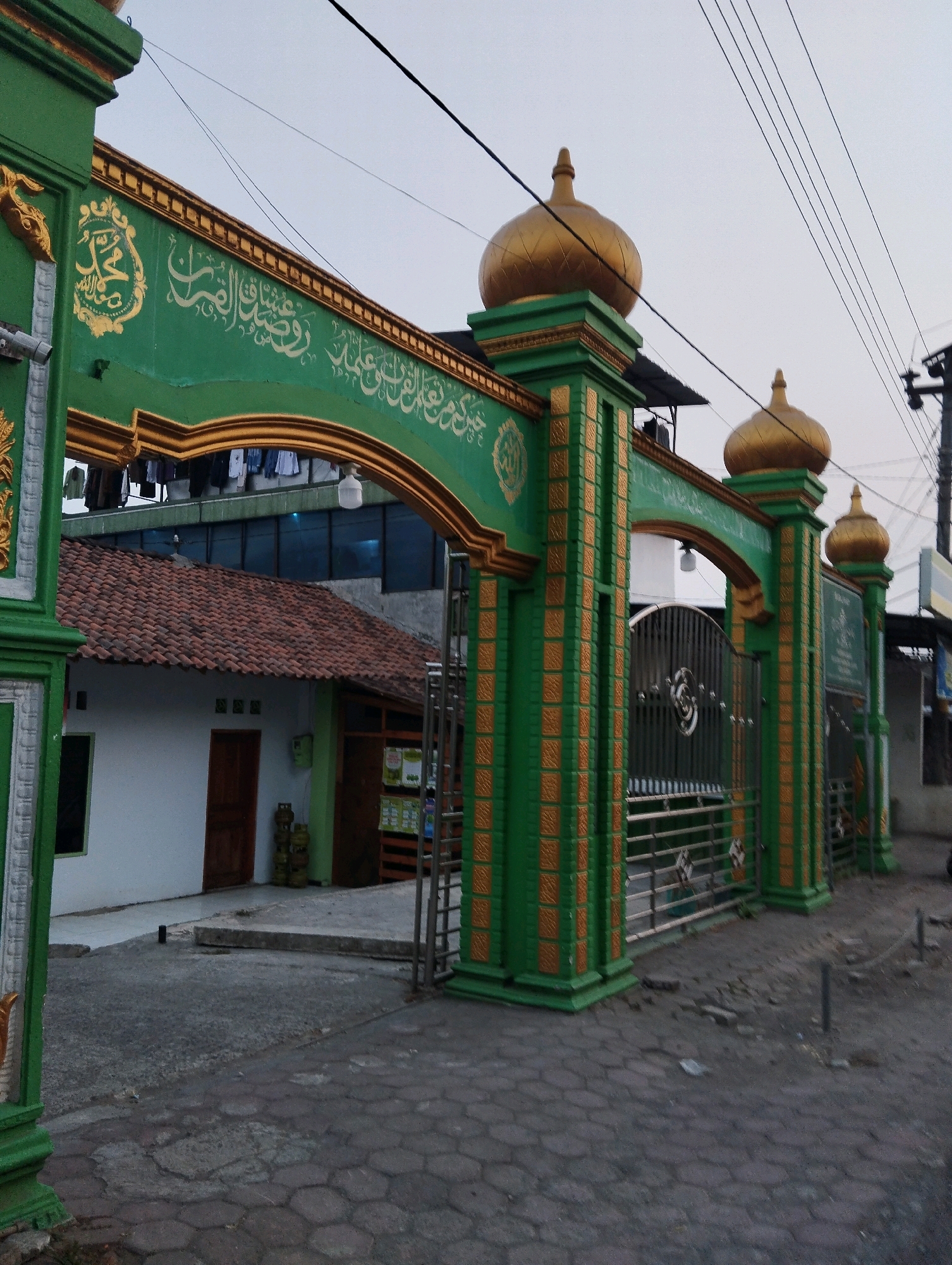 The image shows the ornate green and gold entrance gate of what appears to be a mosque in Salatiga, Indonesia. The gate is decorated with Arabic calligraphy and topped with golden domes. Through the gate, a building with a red-tiled roof and white walls is visible. Several lines of laundry are hanging on a line in front of a darker-colored building behind the mosque entrance. The pavement in front of the gate is made of interlocking hexagonal stones. The sky is overcast, and power lines are visible in the background. No people are visible in the image.