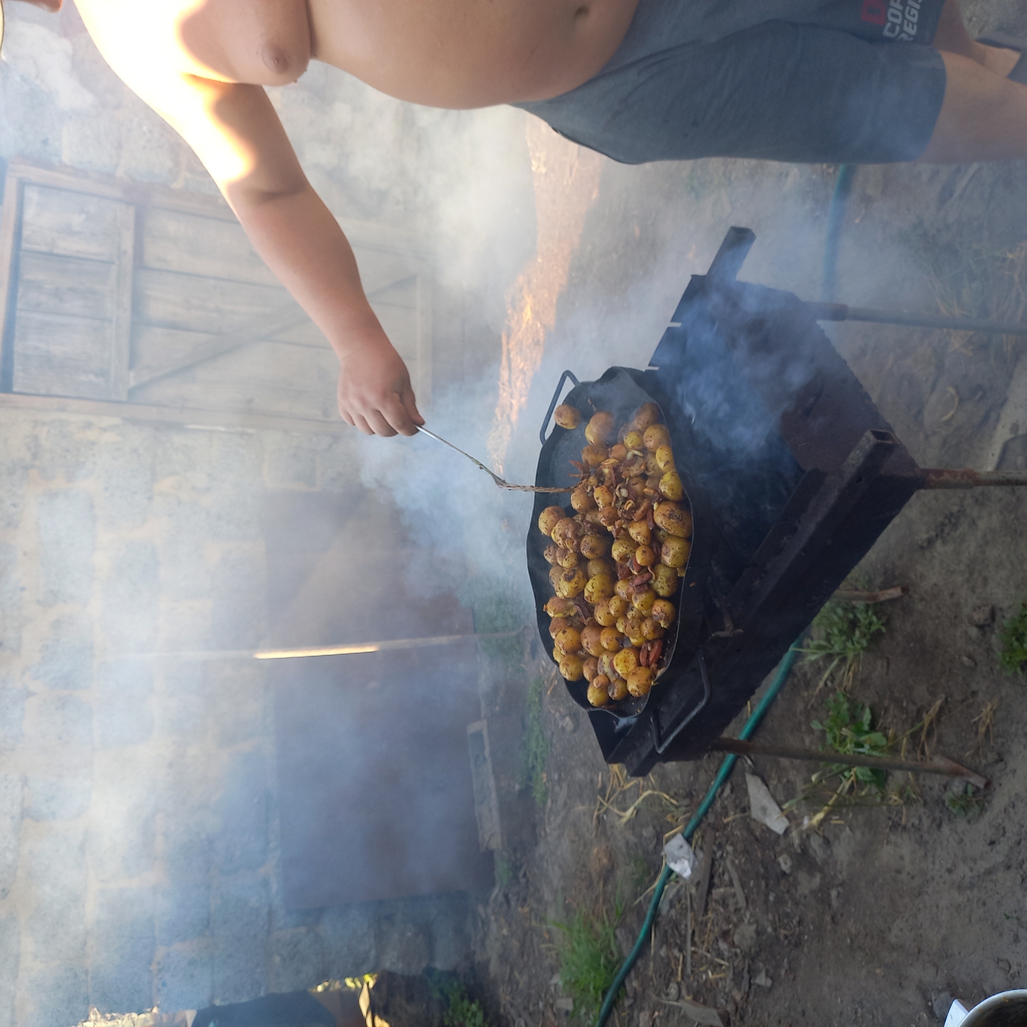 The image depicts an outdoor scene, likely a backyard or rural area, with a focus on a person cooking food. The foreground shows a large, shallow black pan filled with small, golden-brown potatoes, some of which appear to be mixed with pieces of bacon or sausage. The pan is resting on a metal barbecue grill, which is emitting smoke. A person's arm and torso are visible on the right side of the image, wearing grey shorts with a logo that reads "COF REGIS". The person is holding a metal spatula, actively stirring the food in the pan. The background features a rustic stone wall and a wooden door, suggesting a somewhat dilapidated or traditional setting. The ground is covered in dirt, dry grass, and some green vegetation, with a green hose visible to the left of the grill. The overall impression is one of casual outdoor cooking. The location is not specified as Klyntsi, Ukraine, in the provided image or crops.