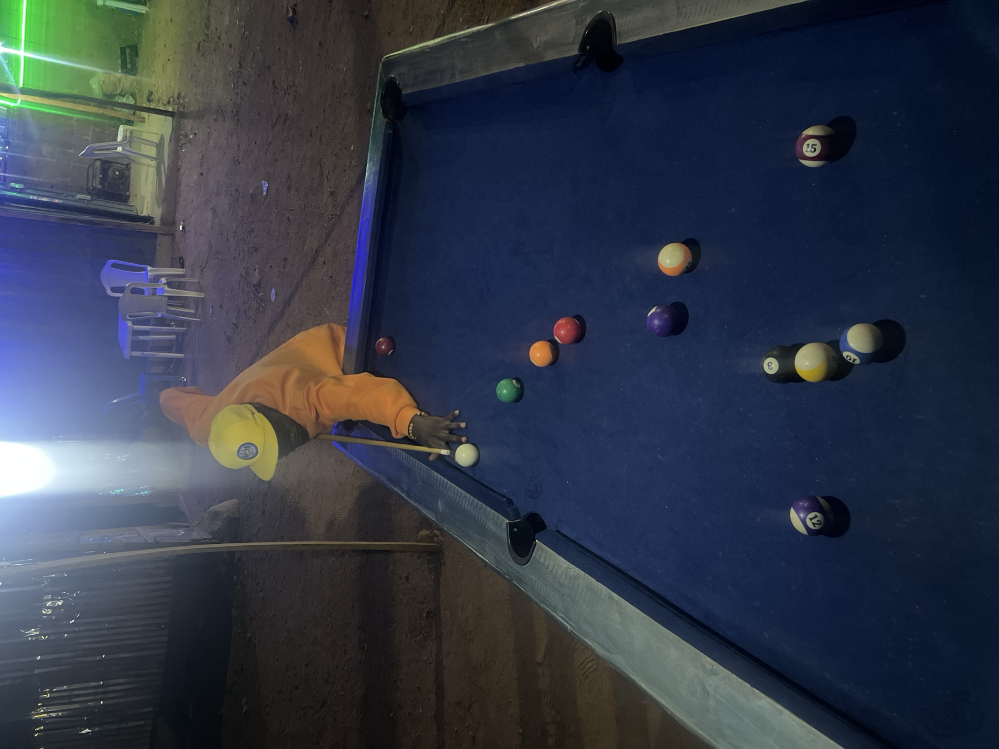 The image depicts a person playing billiards (pool) at night in an outdoor or semi-outdoor setting in Gura Top, Nigeria. The individual, wearing an orange long-sleeved top and a yellow cap, is leaning over a blue-felt pool table, holding a cue stick with their right hand and forming a bridge with their left hand, apparently aiming for a shot.
The pool table is illuminated by artificial lights, which cast shadows from the balls. Scattered across the blue felt are several pool balls, including solid green, orange, and red balls, as well as striped or numbered balls: 15 (purple and white), 12 (purple and white), 10 (blue and white), 8 (black and white), and 3 (black and white). A white cue ball is positioned near the player's cue stick.
The background shows an uneven dirt or gravel ground. Simple white plastic chairs are visible in the mid-ground, and what appears to be corrugated metal sheeting forms a barrier or wall. Bright artificial lights are prominent, creating glare and reflections, particularly on the left side of the frame and on the pool balls. Green light beams are also visible in the upper left background, suggesting a lively, casual atmosphere typical of an evening recreational spot. The time of day is clearly night due to the reliance on artificial lighting.