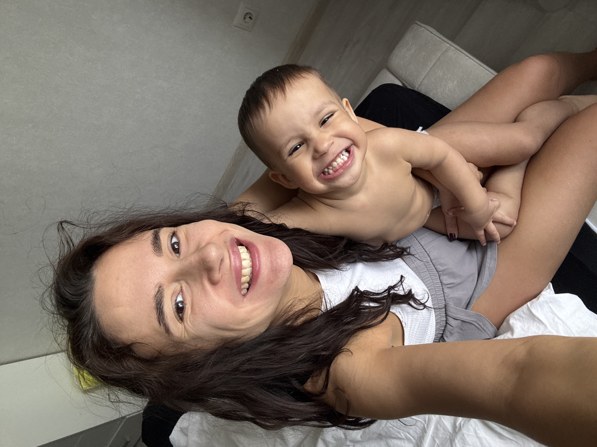 A selfie of a woman lying on a bed or sofa with a baby straddling her lower body. Both are smiling broadly. The woman has dark brown hair and is wearing a white tank top and grey shorts. The baby is shirtless. The room has a neutral color palette, with a white bed sheet, grey wall, and light grey sofa in the background. There is an electrical outlet on the wall. Location is unknown.