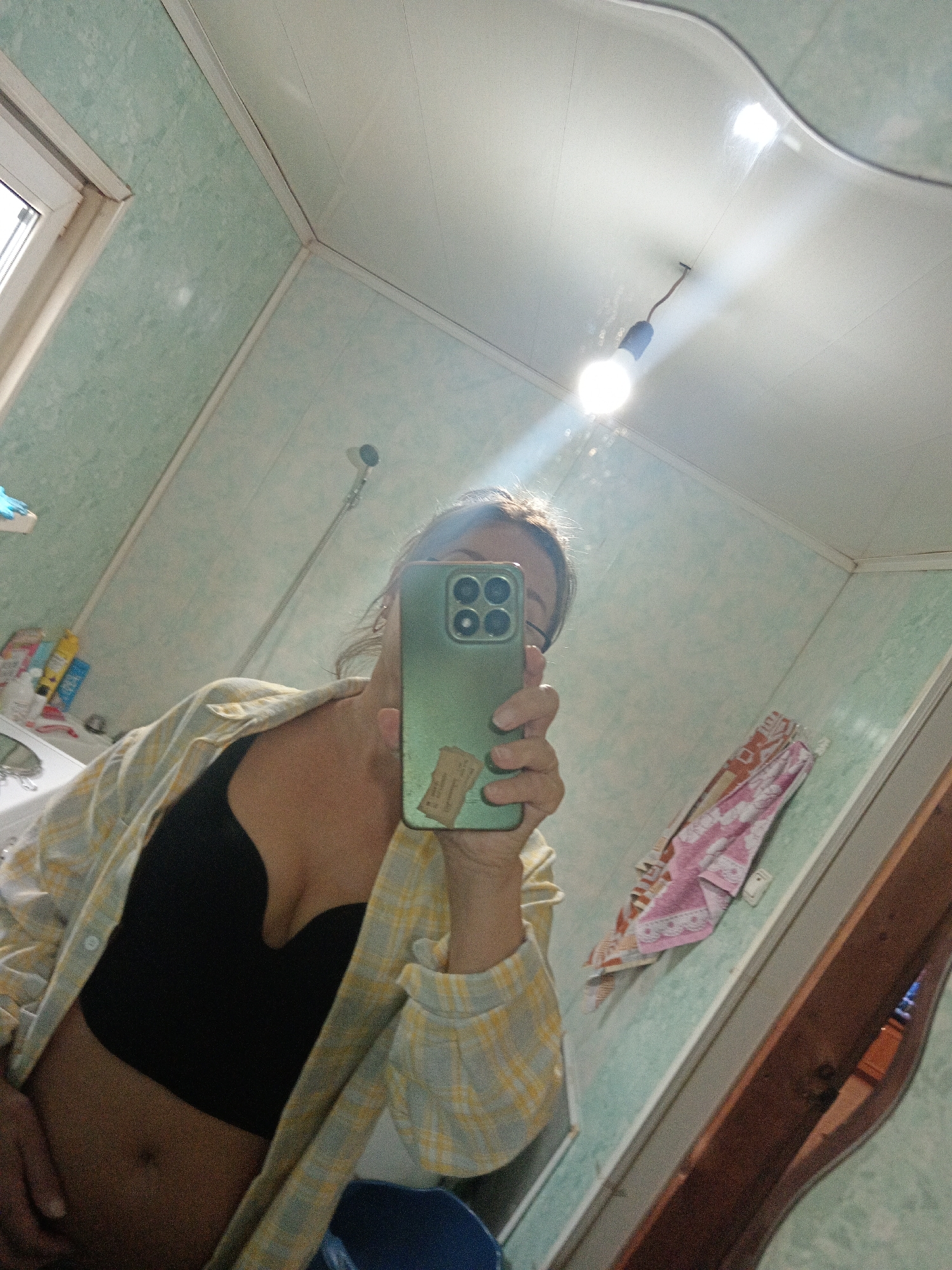 A woman is taking a mirror selfie in a bathroom. She is wearing a yellow and white checkered shirt unbuttoned over a black bra. The bathroom has light green tiled walls and a white ceiling with a single light bulb hanging from it. There is a window on the left side of the image, and a sink with some toiletries visible behind her. A towel is hanging on the wall on the right. The overall impression is of a functional, somewhat dated bathroom. The location of Novopokrovka, Russia is not evident from the image.