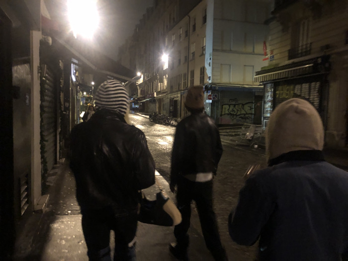 Three individuals walk down a narrow, dimly lit city street at night. The street is wet, reflecting the sparse light. Buildings line the street, their facades marked by graffiti and closed shops. The first person wears a striped beanie, a black leather jacket, and carries a bag. The second, also in a leather jacket and hat, is slightly ahead. The third person, closer to the viewer, wears a blue jacket and a hooded top. The atmosphere is somewhat mysterious and slightly ominous.