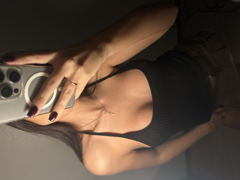 The image displays a mirror selfie of a person, rotated 90 degrees clockwise. The perspective is from the person's upper torso, showing their right hand holding a smartphone up to capture their reflection.
The person is wearing a form-fitting black ribbed tank top or camisole with thin straps. Their skin tone appears warm, with some areas highlighted by an apparent light source, suggesting an indoor setting with directed lighting.
On the ring finger of the hand holding the phone, a simple, thin gold-colored band is visible. The fingernails are painted with a dark red or burgundy nail polish. The smartphone, which has a clear case, appears to be an iPhone model, identifiable by the distinctive triple camera module in the upper left corner of the phone's back and a circular MagSafe-compatible ring on the case.
Around the person's neck, a delicate gold-colored chain necklace with a small cross pendant is visible, resting on their collarbone and décolletage. Dark, straight hair can be seen on the left side of the frame, partially obscuring the person's face (which is not fully visible). On the person's right wrist (visible towards the bottom right of the image if rotated correctly), a thin, light-colored bracelet is worn.
The background behind the person is mostly dark and indistinct, suggesting a dimly lit room or a dark surface behind them. The overall lighting creates a slightly dramatic effect, with highlights on the skin and phone, contrasting with the darker areas of the image. No discernible text is visible on the phone screen or in the background.
