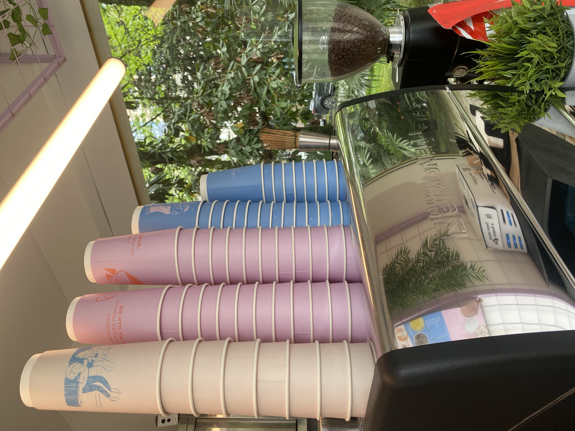 Here is a detailed description of the image:
**General Description:**
The image shows a coffee station, possibly a mobile one, with stacks of paper cups in pastel colors (white, pink, and blue). A coffee grinder sits on top of the station, next to the espresso machine. The shot appears to have been taken in Sochi, Russia, as one of the cups has Russian text printed on it. The station appears to be outdoors, given the view of trees and greenery in the background.
**Specific Objects:**
* **Coffee Cups:** Stacks of paper cups in pastel colors (white, pink, and blue). Some have cartoon drawings on them.
* **Coffee Grinder:** A black coffee grinder with a clear container holding coffee beans.
* **Espresso Machine:** A stainless steel espresso machine, which seems to have the brand name "Simonelli" on it.
* **Brush:** A brush with silver ferrule and wooden handle sits between the cups and the espresso machine.
* **Greenery:** There is artificial plant, next to the coffee grinder.
* **Lighting Fixture:** Above the scene is a bright, cylindrical lighting fixture.
**Environment:**
* The scene appears to be outdoors, with trees and greenery in the background, reflected in the espresso machine.
* The location is likely in Sochi, Russia, based on the text on one of the coffee cups.