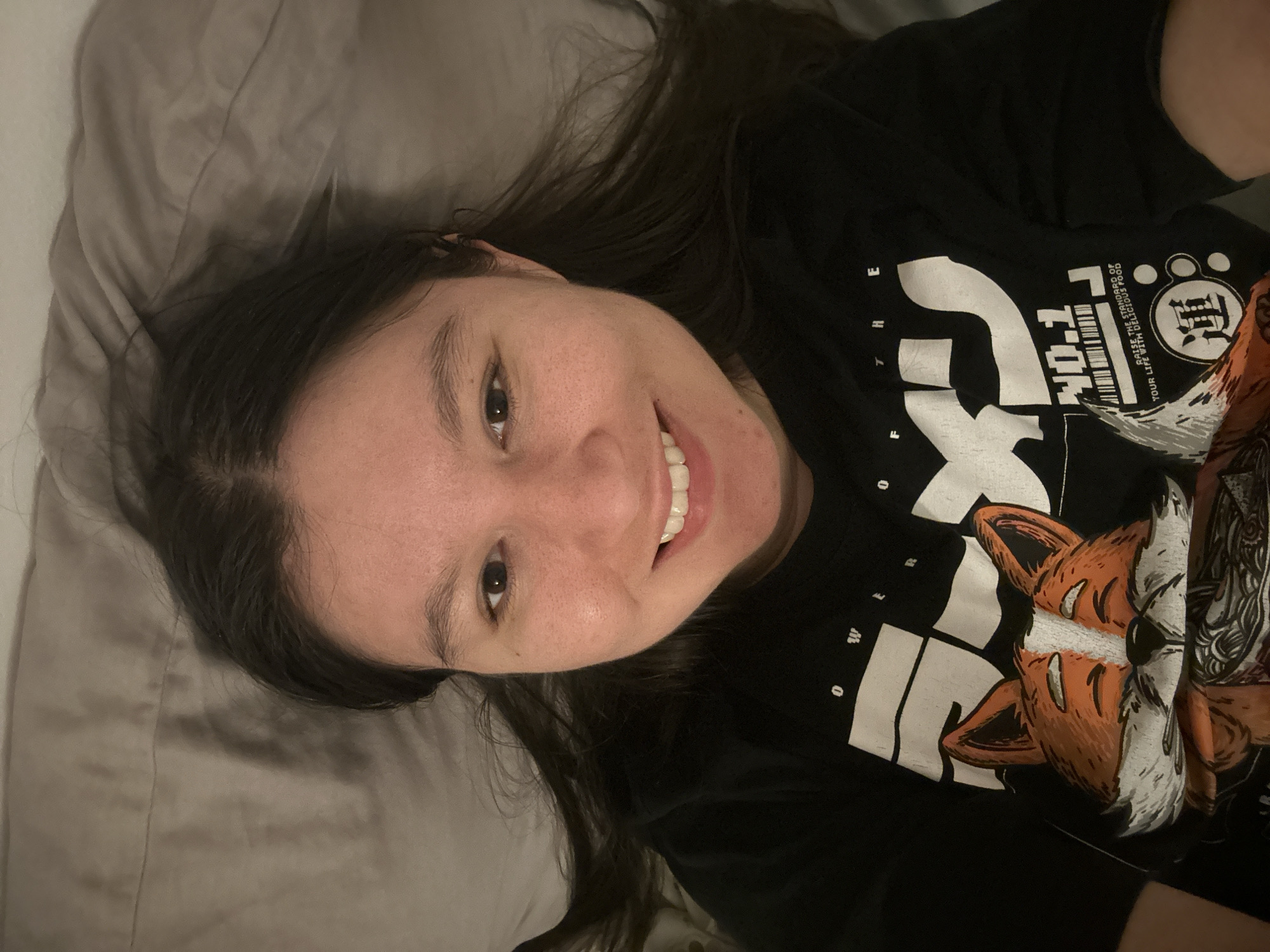 A person is lying on a bed with light gray bedding, facing upward at an angle. They have long dark hair that is partially spread out on the pillow. The person is wearing a black t-shirt with a white and orange design that includes lettering and an image of a fox. The background is out of focus and indistinct. There is no visual indication of the location being Câmara de Lobos, Portugal.