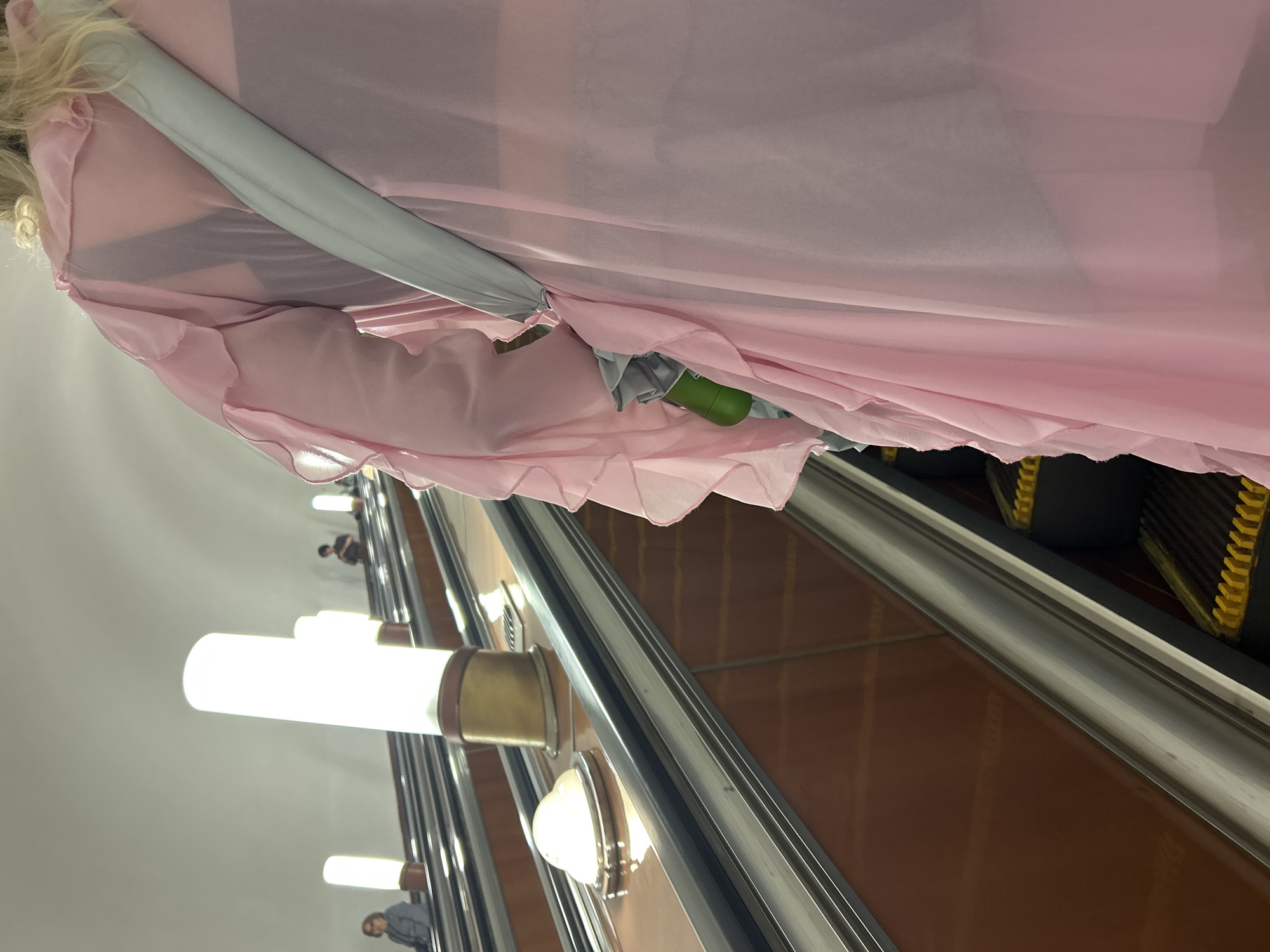 Here is a detailed description of the image:
**Scene Description:**
The image appears to be taken from a low angle inside a metro or subway station, likely an escalator in operation. The focus is on a woman wearing a flowing, light pink garment, partially obscuring the view of the escalator. The escalator has a stainless steel handrail and steps with black rubber traction. Below the handrail is a brown side panel with vertical lines.
**Subjects and Objects:**
* **Woman:** A woman is present, wearing a pink garment. Her blond hair is visible.
* **Escalator:** The escalator is in motion. Details include the handrail, steps, and side panel.
* **Lighting:** There are long, cylindrical light fixtures with brass-colored ends mounted along the escalator shaft. These provide illumination.
* **Umbrella:** A partially visible gray umbrella with a green handle.
* **Other People:** There are a few other people visible in the background on the escalator.
**Actions and Interactions:**
* The woman is likely riding the escalator.
* The escalator is transporting people, including the woman and the others in the background.
**Environment:**
* The setting is an underground metro or subway station.
* The station appears to be relatively clean and well-lit.