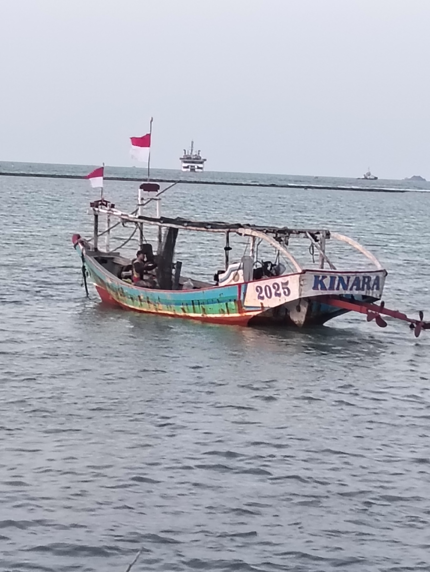 A fishing boat named "KINARA" with the number "2025" painted on its stern is anchored in the waters off Paciran, Indonesia. The boat is colorful, with red, green, and blue accents. Two Indonesian flags are flying from poles on the boat. A person is visible in the boat, and there is a larger vessel in the distance. The water is calm, and the sky is overcast.