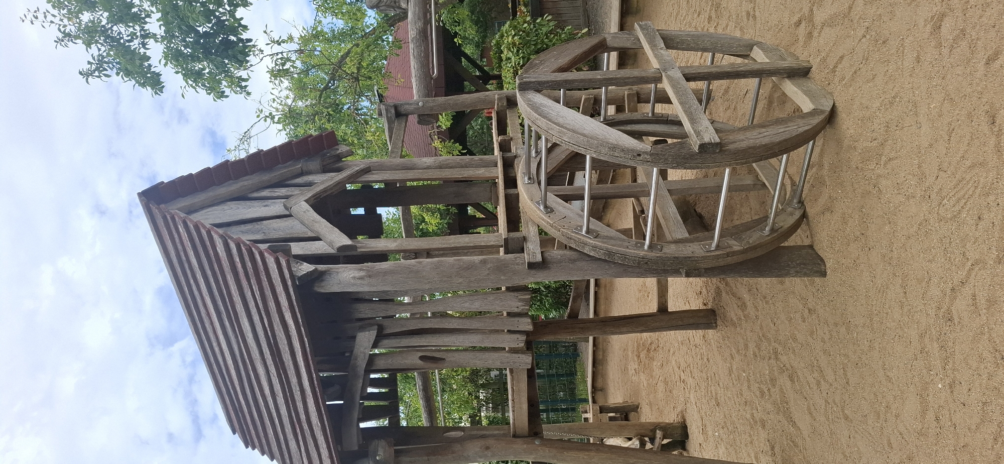 A wooden play structure, resembling an old watermill, stands in a sandy playground. The structure is made of weathered wood and features a pitched roof with red shingles. Two large wooden wheels with metal axles are prominently displayed on the side of the structure. The playground is surrounded by greenery and trees, with a cloudy sky overhead. This playground is located at the Gottfried-Benn-Gymnasium in Hürth, Germany. There are no people visible in the image.