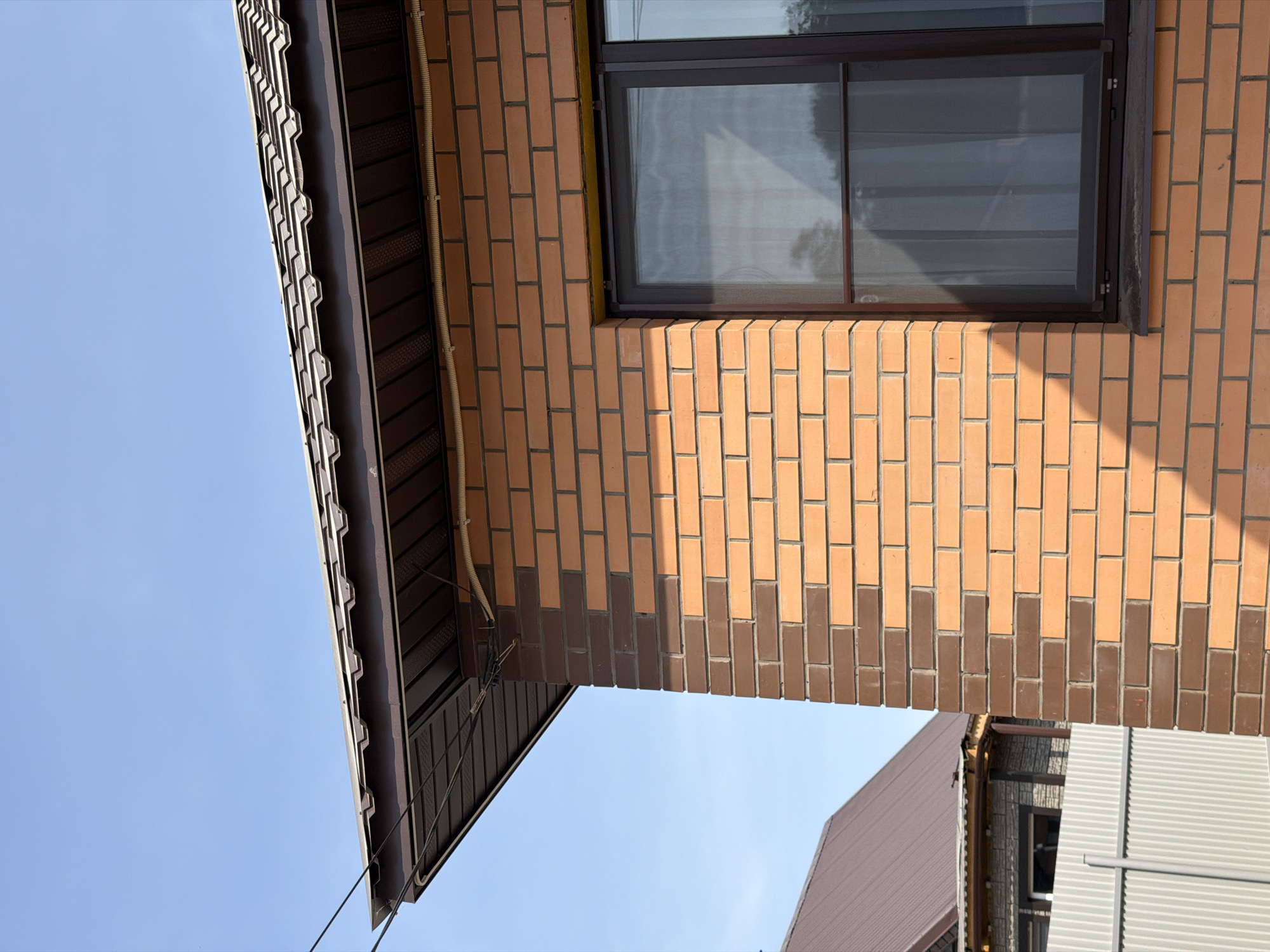 This is an upward-angled view of a building in Chaikovsky, Russia. The building has a brown tiled roof, brick walls, and a dark-framed window. The bricks are a mix of light brown and darker brown. The sky is visible in the background and is clear blue. Another building with a brown roof and grey siding is also visible in the bottom right corner.