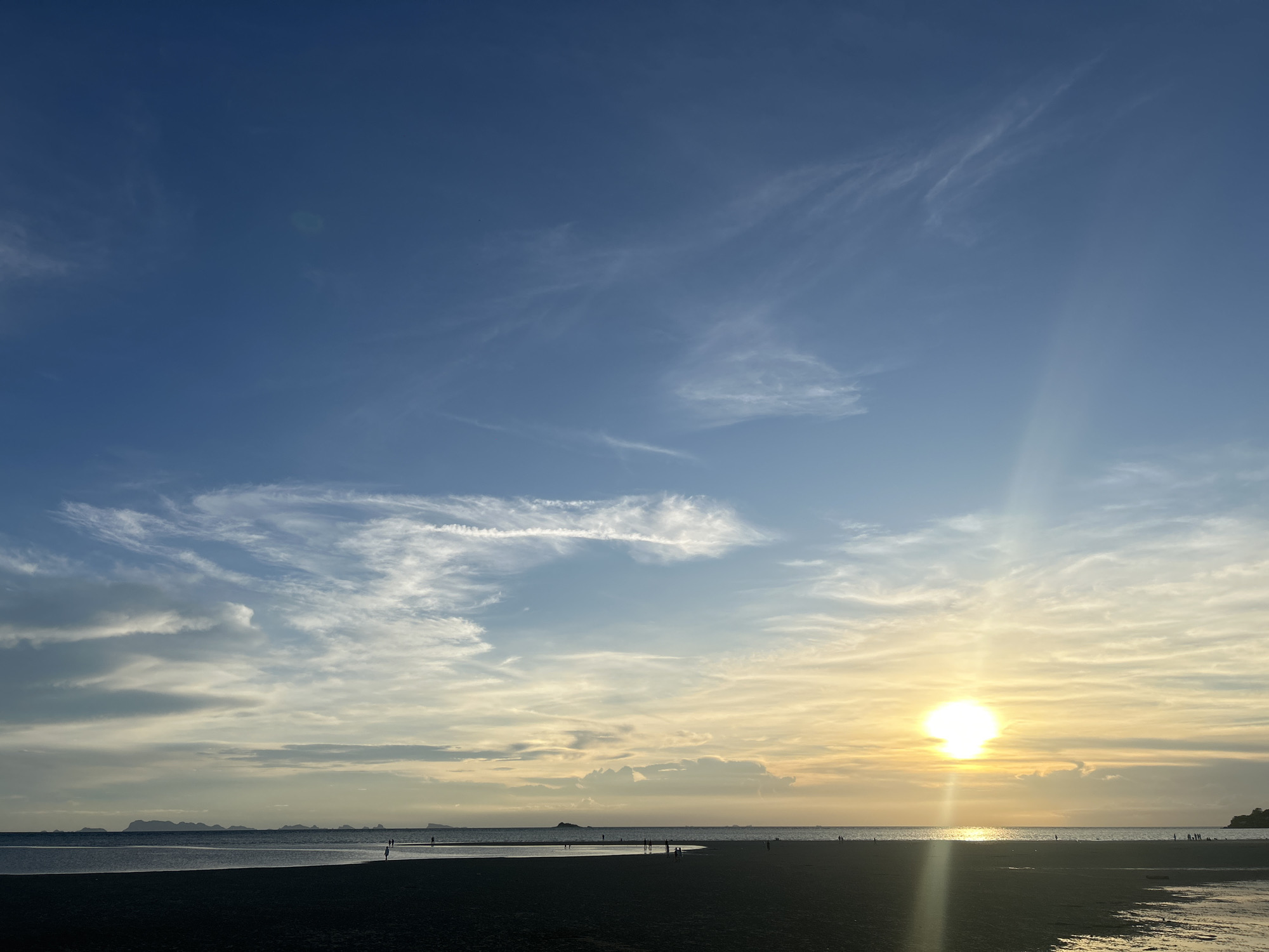 The image shows a sunset scene on a beach in ตำบลเกาะพะงัน, Thailand. The sky transitions from blue at the top to a yellowish-orange near the horizon where the sun is setting. Wispy clouds are scattered throughout the sky. Below the sky is the sea. Some small islands or landmasses are visible on the horizon. The foreground shows a dark sandy beach with a few people scattered along the shore, some standing in shallow water. The sun's reflection can be seen on the wet sand and water, creating a shimmering effect.