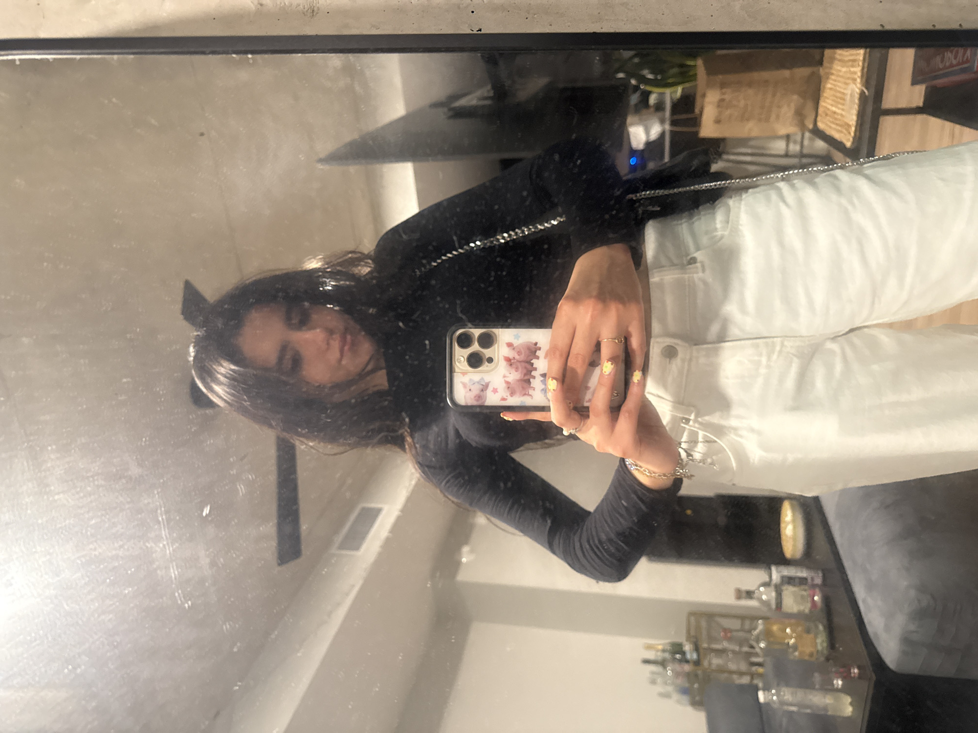 A person with long dark hair is seen in a mirror selfie. The person is wearing a long-sleeved black top and white pants. The phone case has a pattern of cartoon pigs and flowers. The background shows a modern interior setting, including a flat-screen television, a bookshelf, and a bar cart with alcohol bottles. The environment suggests a residential apartment in an urban location, likely in a city such as Austin, United States of America.