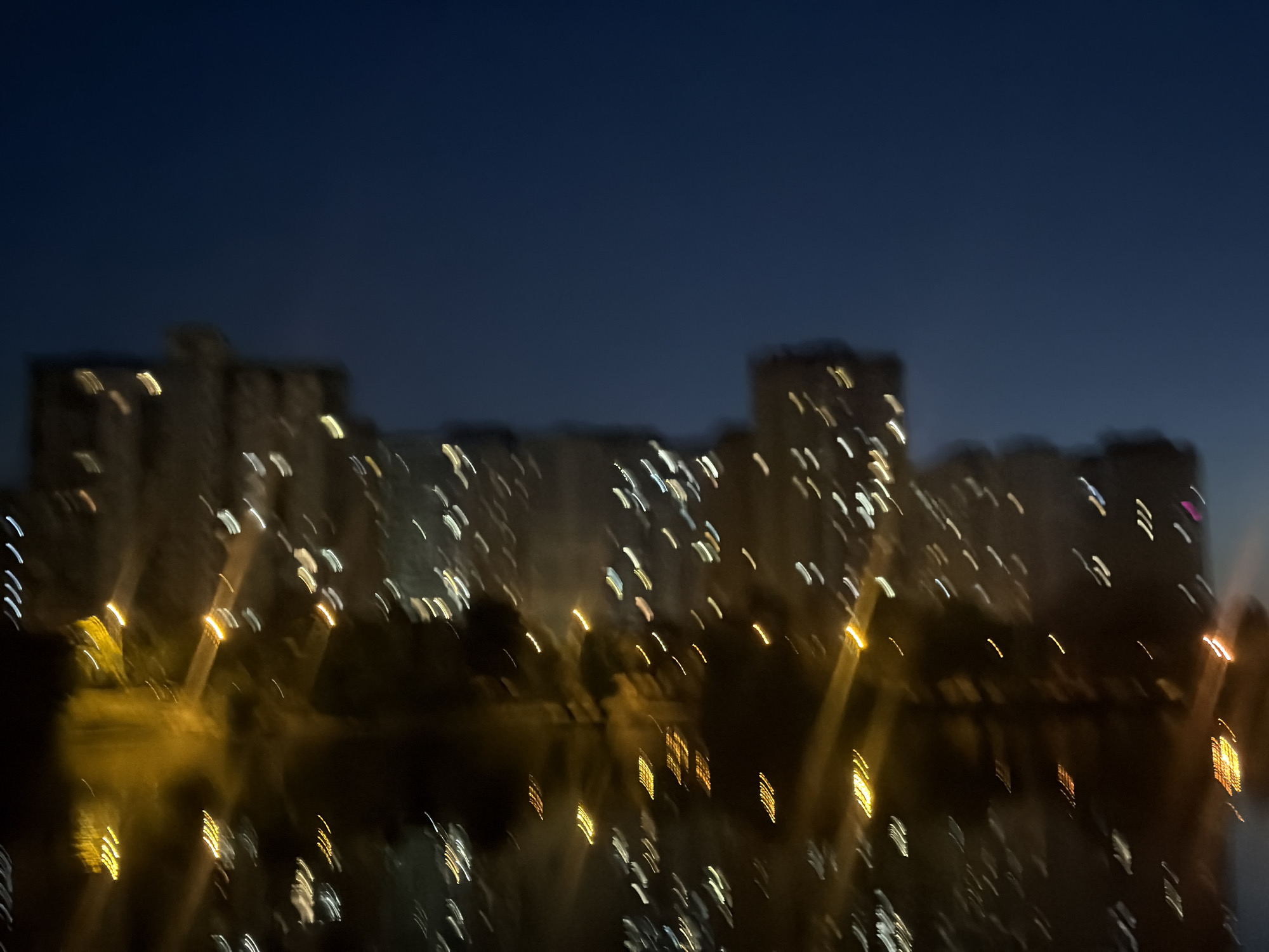 The image captures a blurry, abstract view of a cityscape at dusk or night, likely in Kyiv, Ukraine. The dark blue sky dominates the upper portion, with a dark horizon line featuring indistinct, elongated streaks of light that suggest buildings or city lights. These lights are rendered as elongated blurs, indicating motion blur or a long exposure. Below the horizon, a body of dark water reflects the lights, creating a shimmering effect with more elongated streaks. The overall impression is one of a city illuminated by numerous lights, perhaps from apartment buildings or streetlights, seen through the effect of motion or a low-light photography technique. The image is devoid of any discernible people or specific recognizable landmarks.