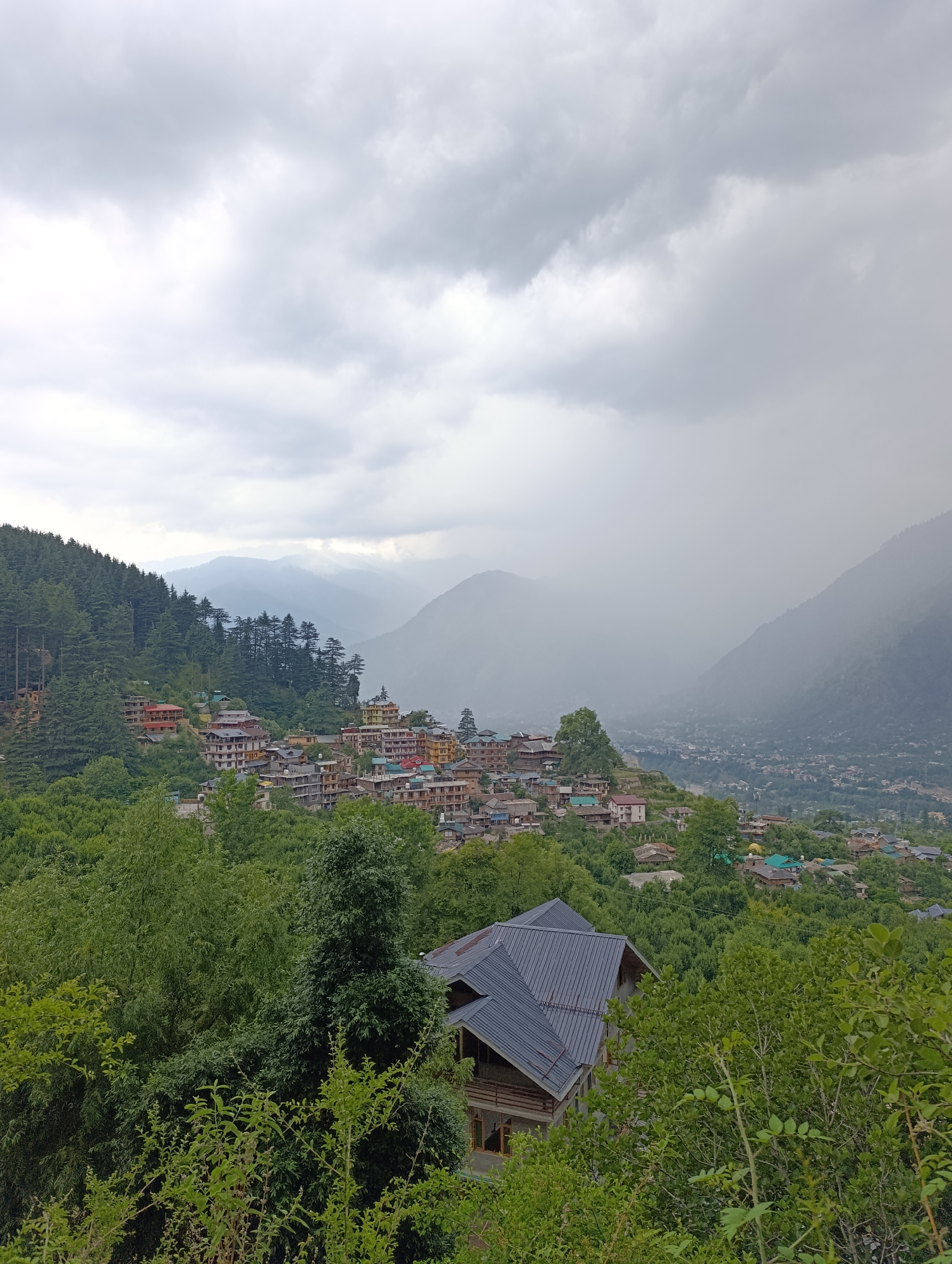 This panoramic image captures a sprawling mountain village or town, identified as Naggar, India, nestled within a lush, forested landscape under a heavily overcast sky. The scene is dominated by shades of green and grey, reflecting the abundant vegetation and cloudy weather.
In the foreground, dense green foliage and the tops of deciduous and coniferous trees fill the lower portion of the frame. Partially visible on the bottom left, a house with a distinctive dark grey, multi-faceted sloped roof is tucked amongst the trees, suggesting a modern architectural style in harmony with the natural surroundings.
The midground showcases the village, featuring numerous buildings built into the hillside. These structures display varied architecture, with some featuring traditional sloped roofs and others appearing more contemporary. They are clustered together, interspersed with tall green trees, creating a mosaic of human settlement within the natural environment. The buildings are multi-storied and appear vibrant against the green backdrop.
In the background, successive layers of mountains recede into the distance, their forms softened by atmospheric haze. The sky is entirely covered with thick, grey clouds, indicating overcast weather. A notable detail is the visible rain or mist falling over the distant valley and mountains on the right side of the image, creating a blurred, ethereal effect and further obscuring the far-off landscape. A larger urban area can be glimpsed on the valley floor in the far distance, partially shrouded by the weather conditions.
The time of day appears to be daytime, but the heavy cloud cover gives the scene a diffused, soft light, typical of a rainy or pre-rainy day. There are no people visible, nor are there any discernible activities taking place. No text is visible within the image. The overall impression is one of peaceful, robust mountain life amidst the dramatic elements of nature.