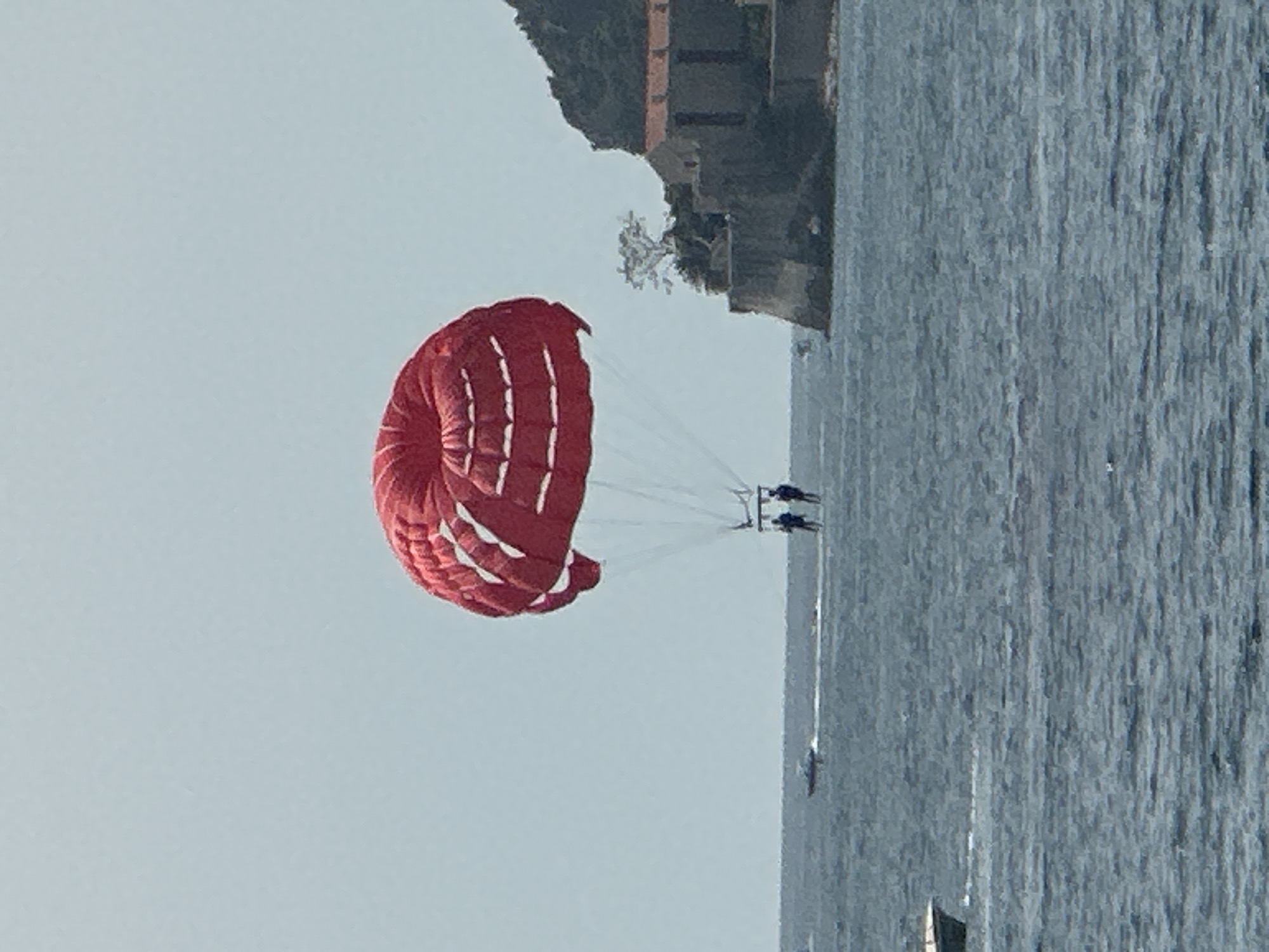 The image depicts a bright, sunny day at sea, with a prominent red parachute carrying two individuals parasailing in the mid-ground. The parachute is large and circular, with white horizontal stripes. Below the parachute, two people are visible, suspended by harness lines. In the background, a rugged, rocky coastline is visible, featuring a small cluster of stone buildings with terracotta roofs, likely an ancient settlement. A single, sparse tree grows on the cliffside. The sea is a deep blue-grey, with gentle ripples indicating calm conditions. Several small boats are scattered across the water, including one in the far left, which is partially submerged and appears to be a small speedboat. Further in the distance, another boat is seen leaving a wake on the water. The sky is a pale, hazy blue, suggesting a warm, clear day. The overall scene captures a recreational activity taking place in a picturesque coastal setting, potentially near Budva, Montenegro, given the architectural style of the buildings and the rugged coastline.