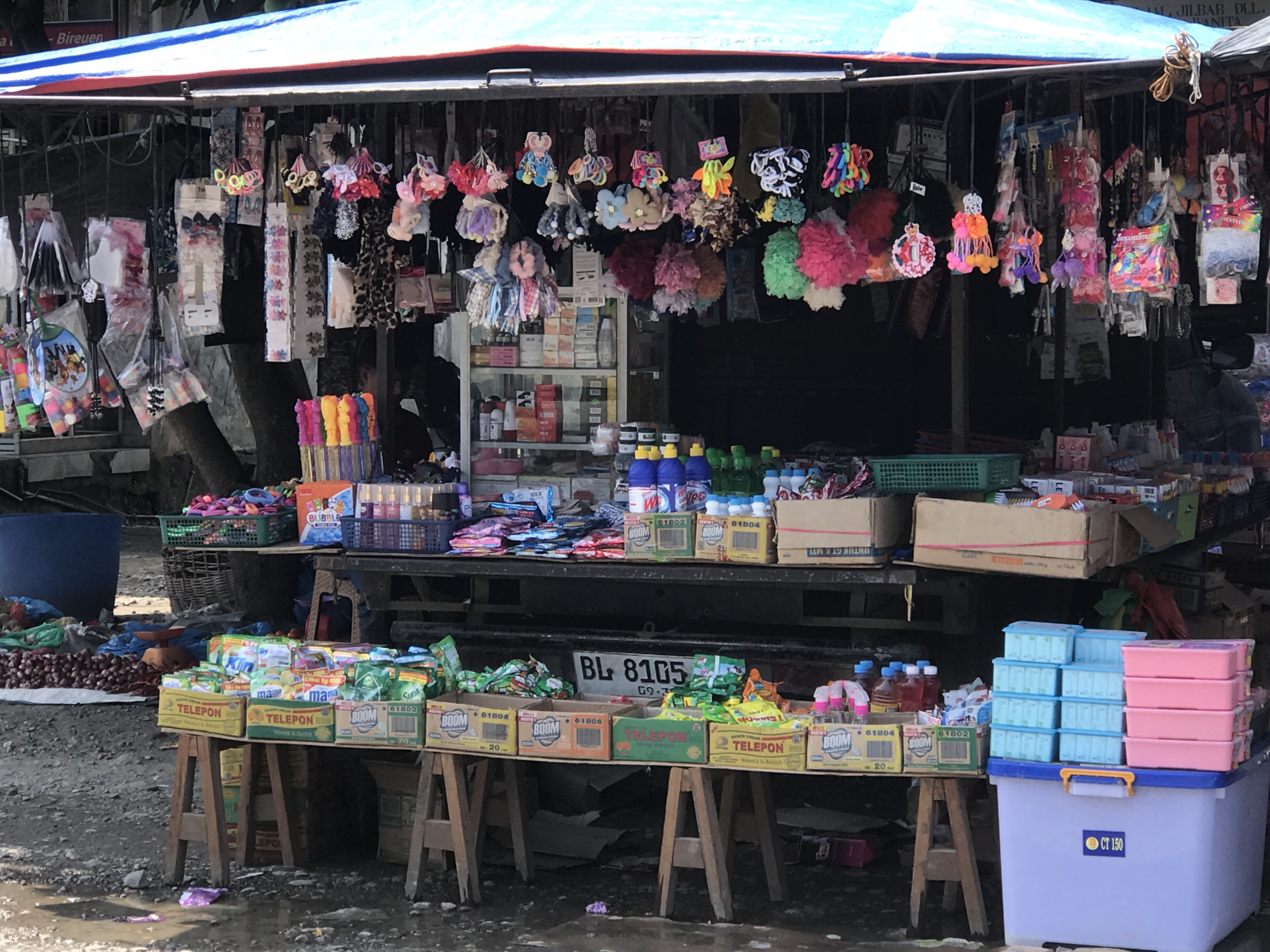 This image depicts a roadside vendor stall located in Tingkeummanyang, Indonesia. The stall is sheltered by a blue tarp and constructed of wooden tables, upon which various goods are displayed. Hanging from the stall's frame are numerous hair accessories, including scrunchies and headbands. Shelves behind the hanging items showcase additional merchandise such as toiletries, household goods, and cosmetics. The foreground features boxes of powdered detergents labeled "BOOM" and "TELEPON", alongside stacks of plastic containers. In the background, a cluster of onions is visible near a blue bucket. The license plate "BL 8105" is partially visible. The ground is uneven and appears wet.