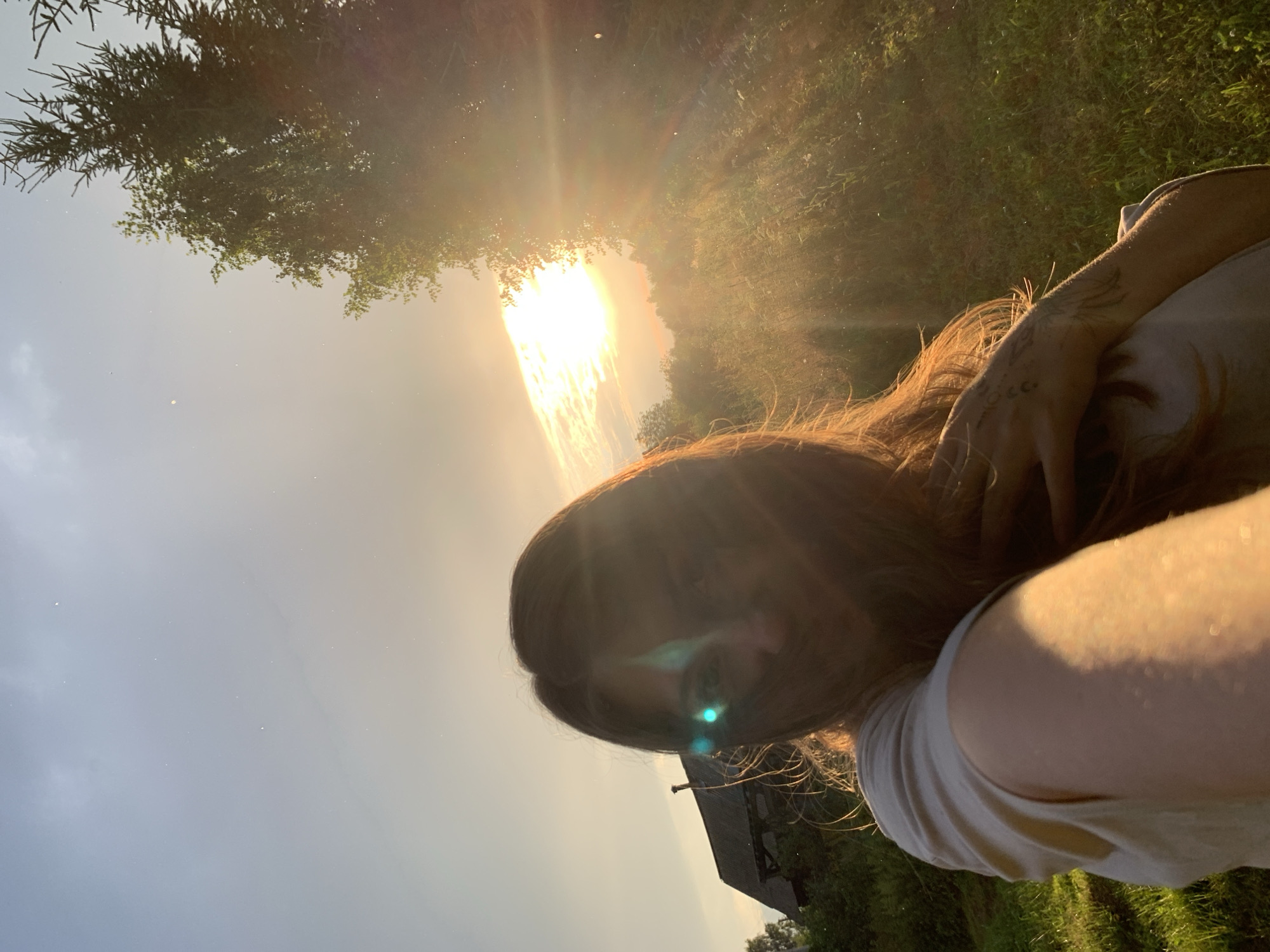 The image is a selfie taken outdoors, likely in a rural setting in Zasechnyi, Russia, during sunset given the warm, golden light. The sun is low in the sky, casting strong rays that create lens flare and illuminate dust motes in the air. The foreground is dominated by a person with long, reddish-brown hair, who is holding their arm across their face, obscuring part of it. The person is wearing a light-colored t-shirt. Visible tattoos on the person's arm include a wing and a crescent moon with a dot.
The background shows a grassy field bathed in the sunset light, with some trees or foliage to the left and right. In the distance, a dark, rustic building, possibly a barn or shed with a steeply pitched roof, is partially visible amongst the trees. The sky is a bright, hazy orange and yellow near the sun, transitioning to a lighter, possibly blue or grey, tone further away. The overall mood appears relaxed and contemplative, possibly reflecting on the end of the day. There is no visible text in the image.