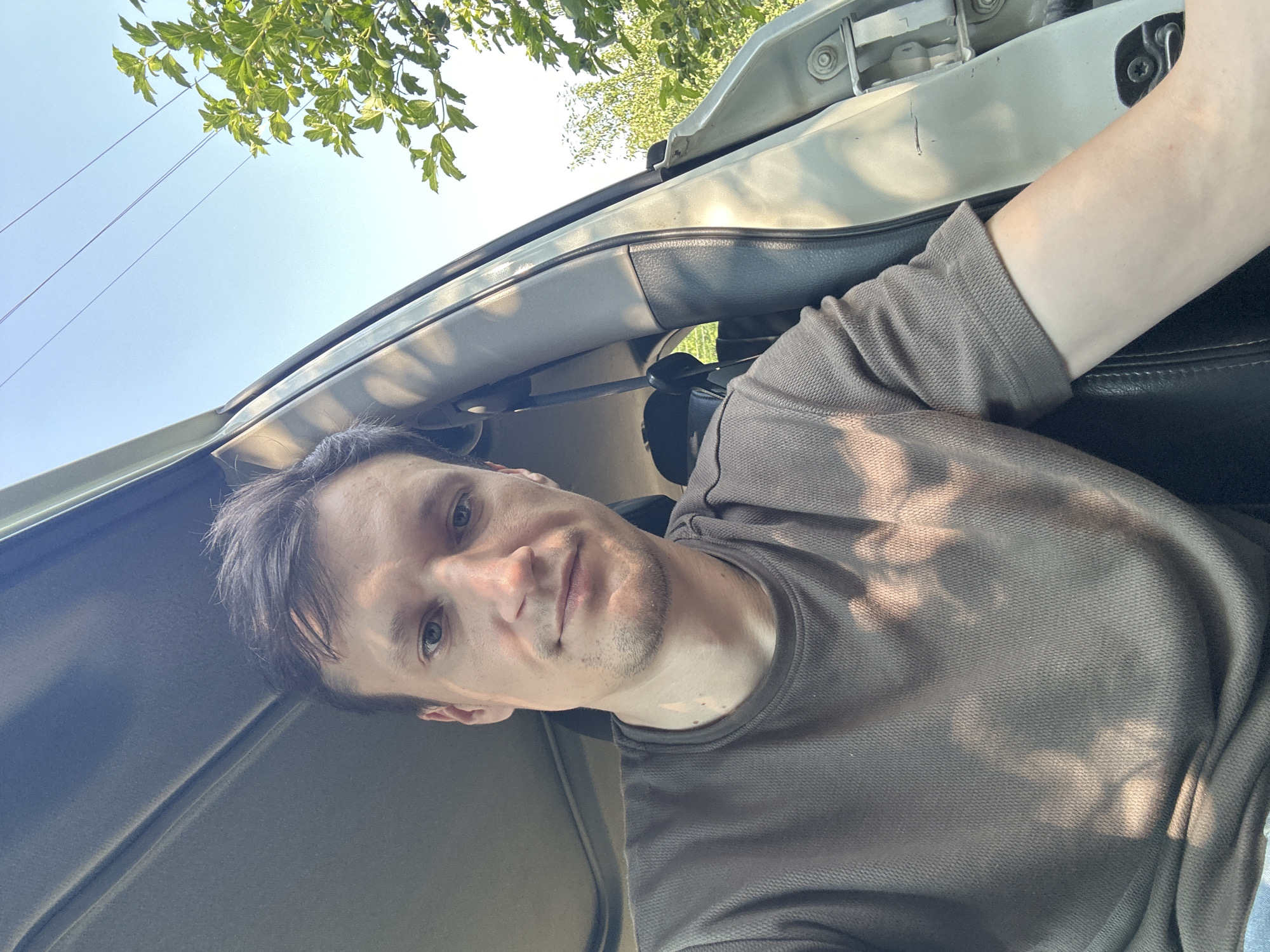 A man with short brown hair and light blue eyes is sitting in the driver's seat of a car, taking a selfie. He is wearing a brown, waffle-knit t-shirt and has a slight stubble on his face. The car's interior is visible, with the door ajar, revealing a portion of the seat and the car's frame. Sunlight streams in, casting shadows on the man's shirt and the car's interior. Outside the car, green foliage and a clear blue sky are visible. The scene appears to be taking place outdoors, possibly in a rural area or a park, given the presence of trees and the natural light. The image does not contain specific landmarks or details that would allow for a precise location within Shatava, Ukraine, beyond the general context of being in that region.
