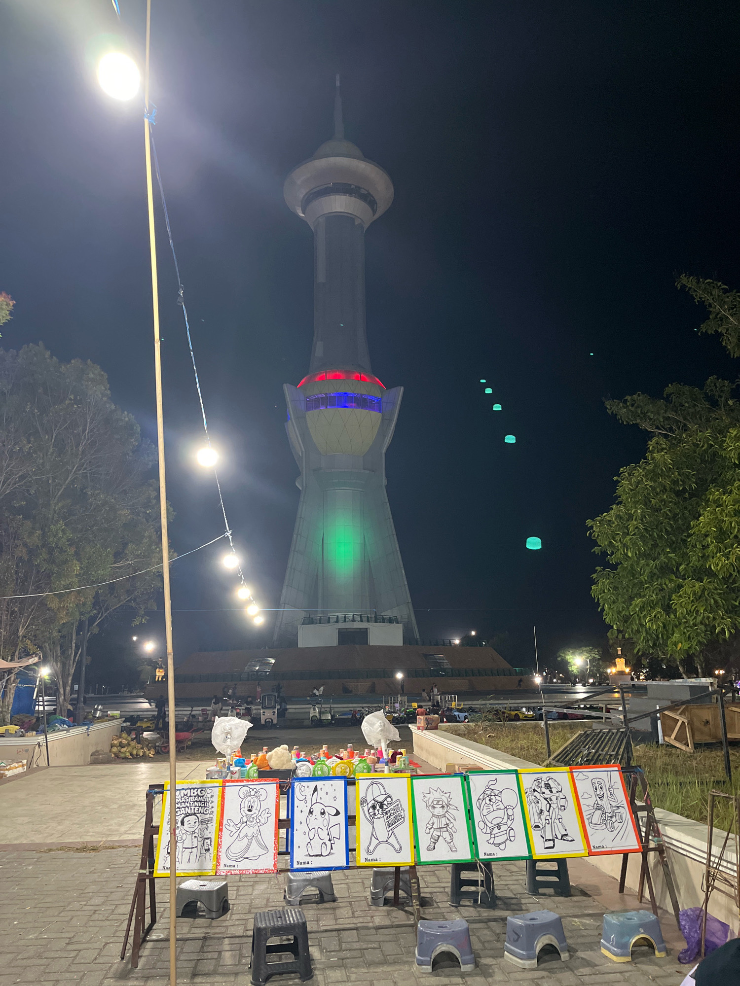The image captures a nighttime scene at the Kendari City Tourism Object (TTS), also known as the Kendari Dragon Tower. The iconic tower, illuminated with green and red lights, dominates the background. In the foreground, a row of coloring pages featuring cartoon characters like Mickey Mouse, Pikachu, Naruto, Doraemon, and a robot is displayed on a stand, with small stools placed in front. String lights are visible overhead, and trees line the left side of the image. Several people are seen in the middle ground, some near stalls and others near what appear to be amusement rides. The area is paved, suggesting it is a public space. The overall atmosphere is that of a lively night market or festival setting.