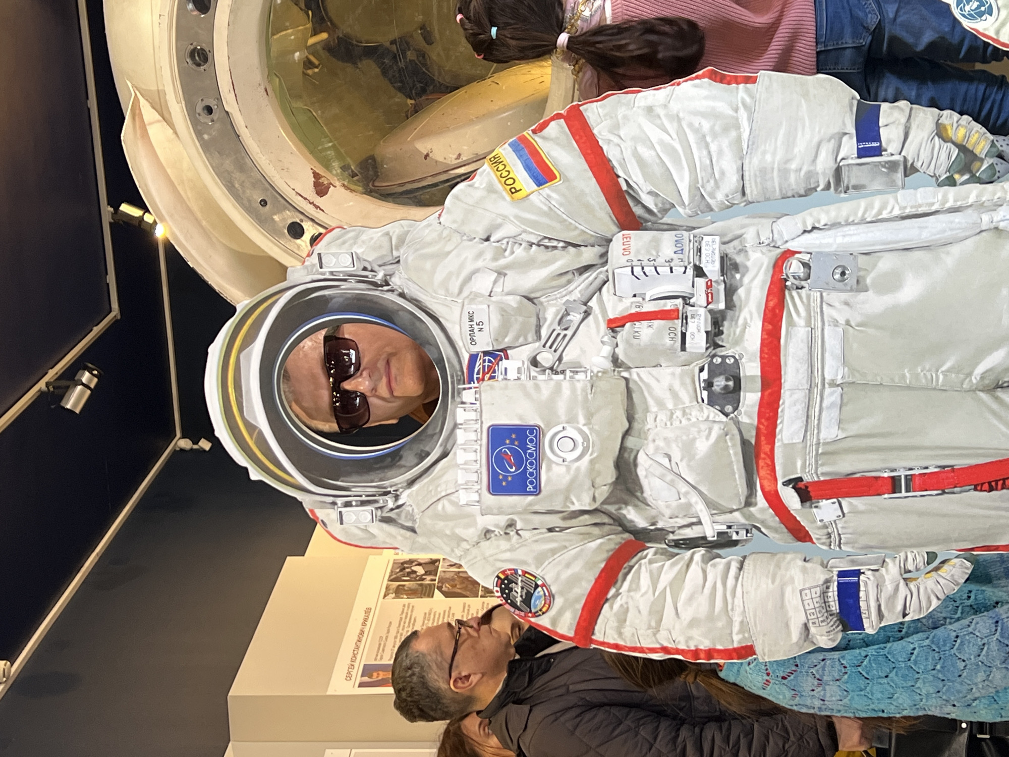 A medium indoor shot shows an exhibit of a Russian astronaut suit, presumably in Saint Petersburg, Russia. A man wearing sunglasses is positioned in the astronaut suit cutout. A woman in a pink sweater and jeans stands to the right of the suit. A man in a dark jacket stands to the left of the suit, in front of a display titled "SERGEI KONSTANTINOVICH." Behind the suit is a section of a space capsule. The suit is labeled with a Russian flag and the word "Россия" and "РОСКОСМОС" and "ОРЛАН МКС N5". The text on the suit, in Russian, seems to relate to readings and settings. The scene is well-lit, showcasing the exhibit.