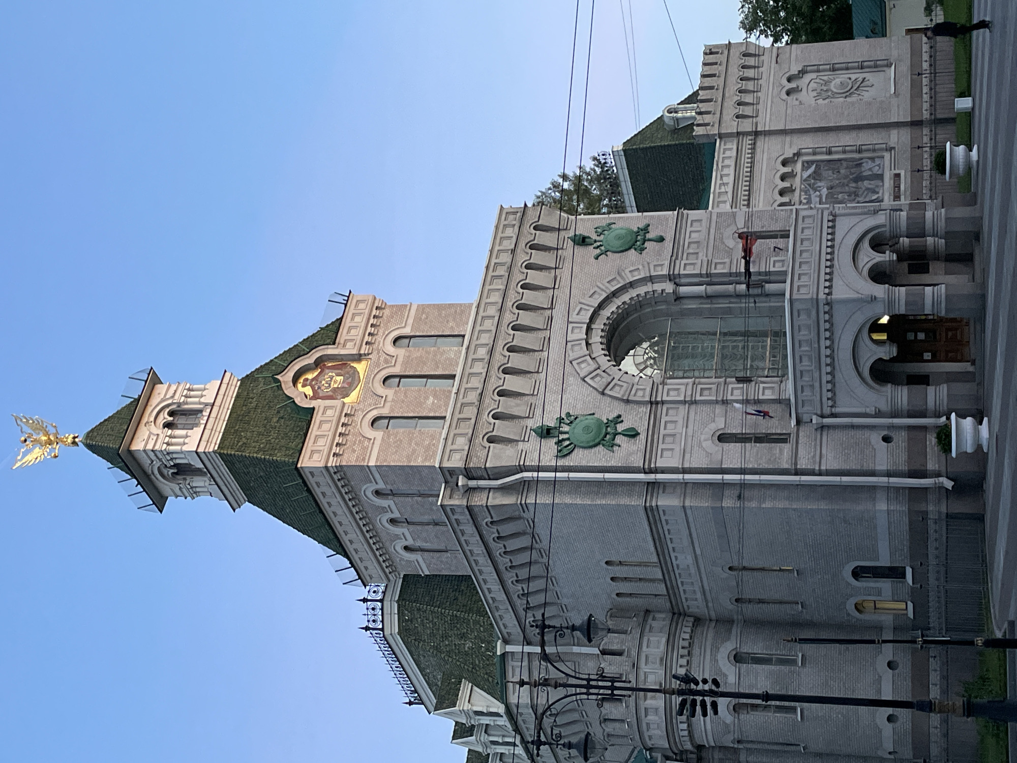 The image shows the Tauride Palace in Saint Petersburg, Russia, from a low angle on a clear day. The palace is a grand neoclassical building with a central tower topped by a golden double-headed eagle. The tower has a green roof and arched windows. The walls are made of light-colored brick, with decorative green crests on the facade. There is a large arched window above the entrance. In the foreground, there is a lamppost with multiple lights and a person in dark clothing standing to the right of the palace. The sky is a clear blue.