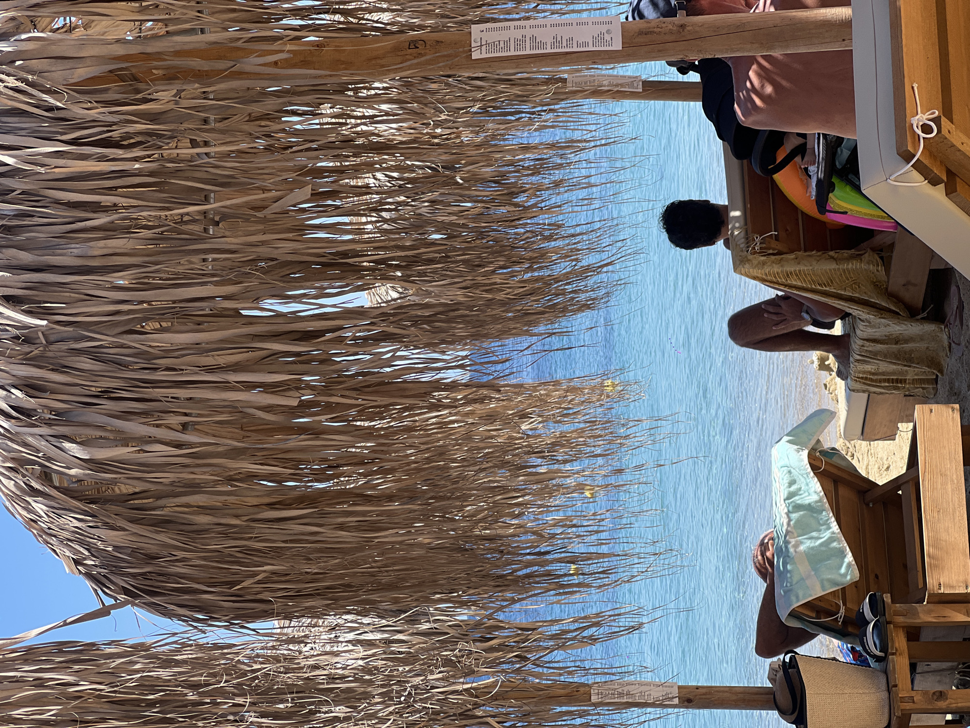 The image captures a serene beach scene in Achladias, Greece, under the shade of palm-thatched umbrellas. The turquoise waters of the Aegean Sea stretch out towards the horizon, meeting a clear blue sky. Several individuals are relaxing on lounge chairs. In the foreground, a person is reclined on a wooden lounger, partially obscured by a light blue and white striped towel. To the left, a straw bag and a pair of flip-flops rest on a wooden table. Further back, two more individuals are seated on lounge chairs. One person, facing away from the camera, is leaning back and looking out at the sea. Beside them, a baby in a stroller is visible, with colorful inflatables nearby. The scene is one of peaceful leisure, with the gentle sound of the waves and the warmth of the sun implied.