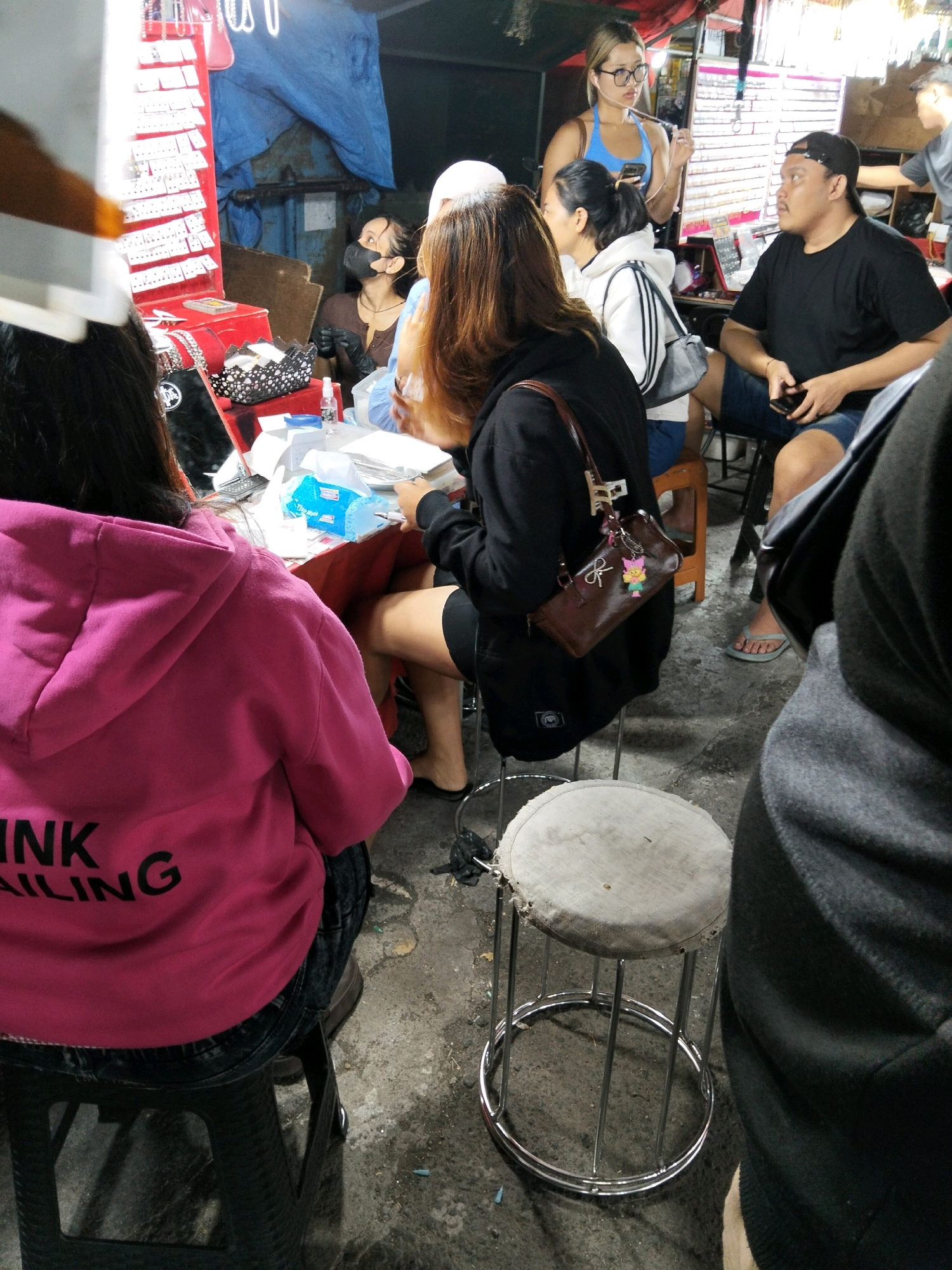 A group of people are gathered around a stall in a street market in Denpasar, Indonesia. The stall appears to be selling jewelry, with various items displayed on a red rack. Several individuals are seated on stools, while others are standing. One woman, wearing a bright pink hoodie, is seated in the foreground. Another woman, in a black hoodie, is seated to her left. A man in a black t-shirt and shorts is seated to the right, looking towards the stall. A woman in a blue top is standing behind the stall, holding a phone. The environment is dimly lit, suggesting it might be evening or the stall is covered. The ground is concrete, typical of street market areas.